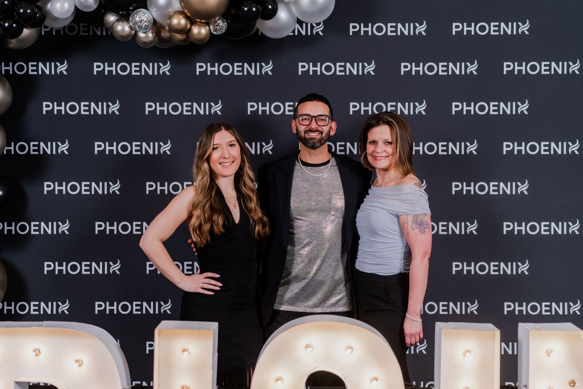 Three people stand smiling in front of a black backdrop with a repeating 