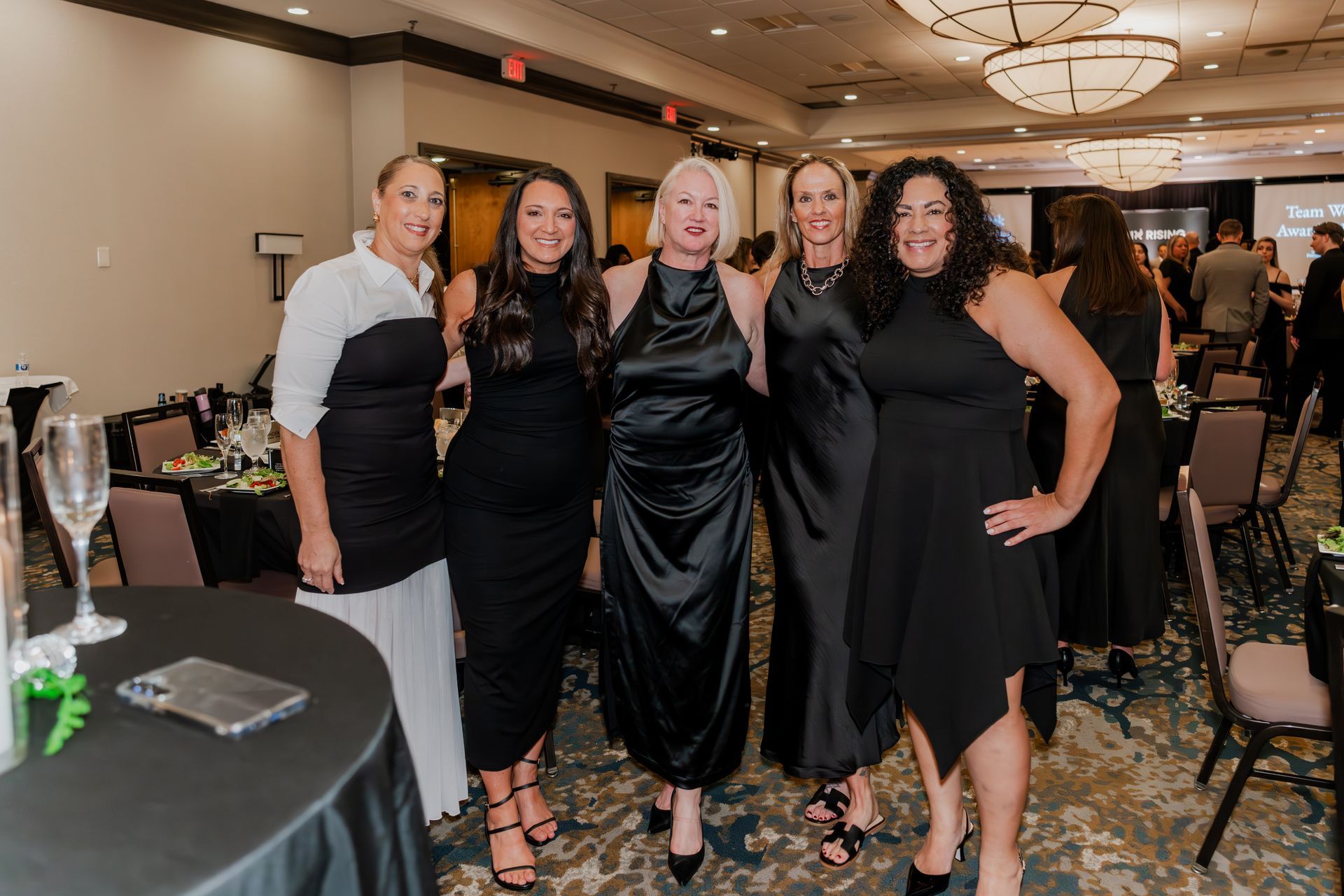 Five smiling people in formal black and white attire pose for a photo at a banquet hall event.
