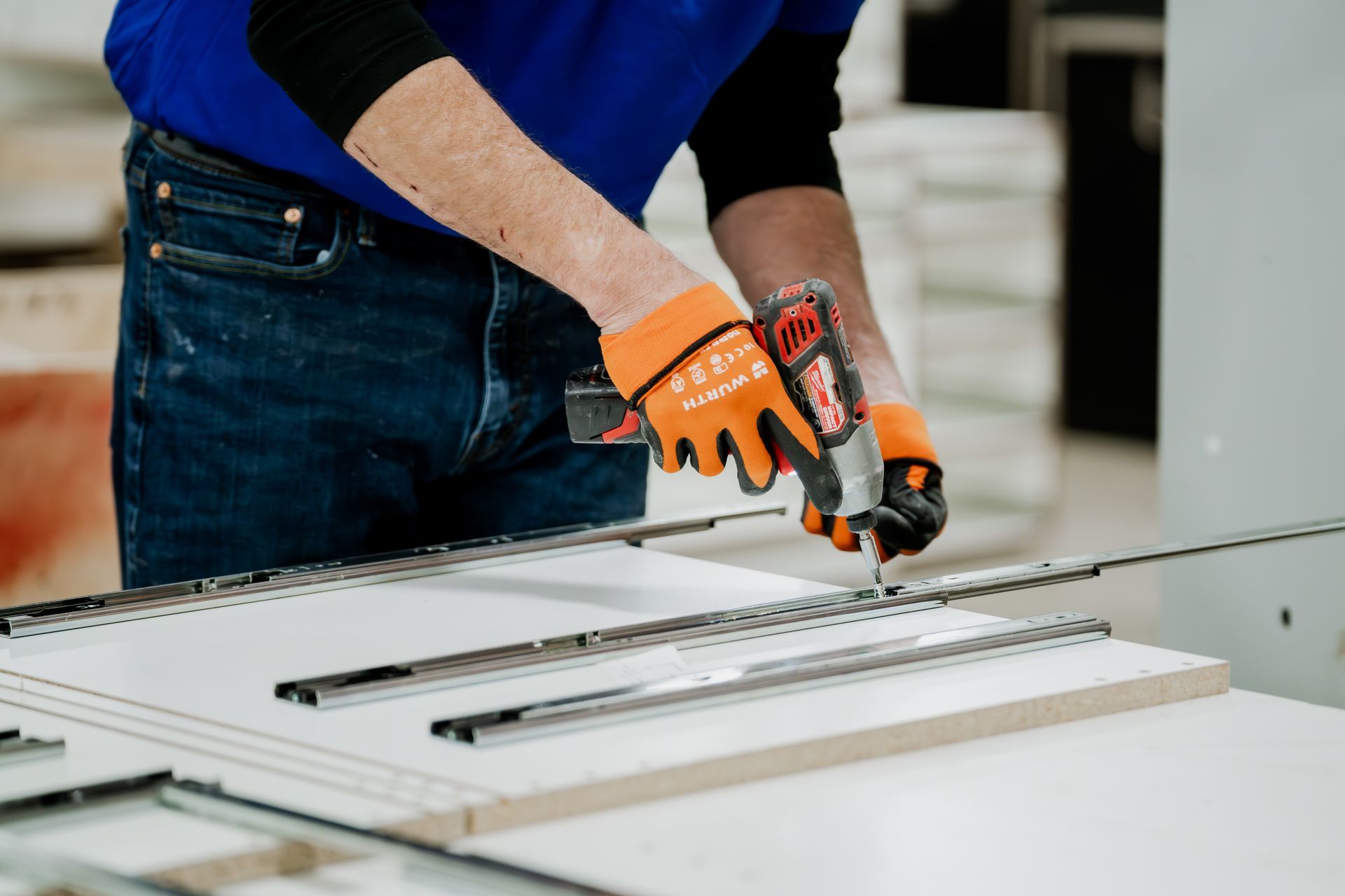 Person using a power drill on a metal track, wearing gloves and a blue shirt in a workshop.