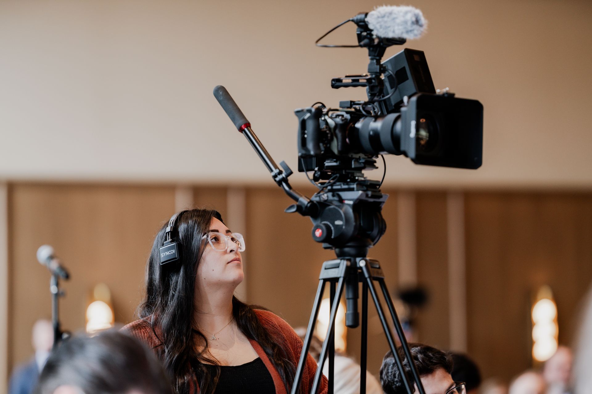 Woman adjusting a video camera on a tripod, microphone attached. Indoor setting, soft lighting.