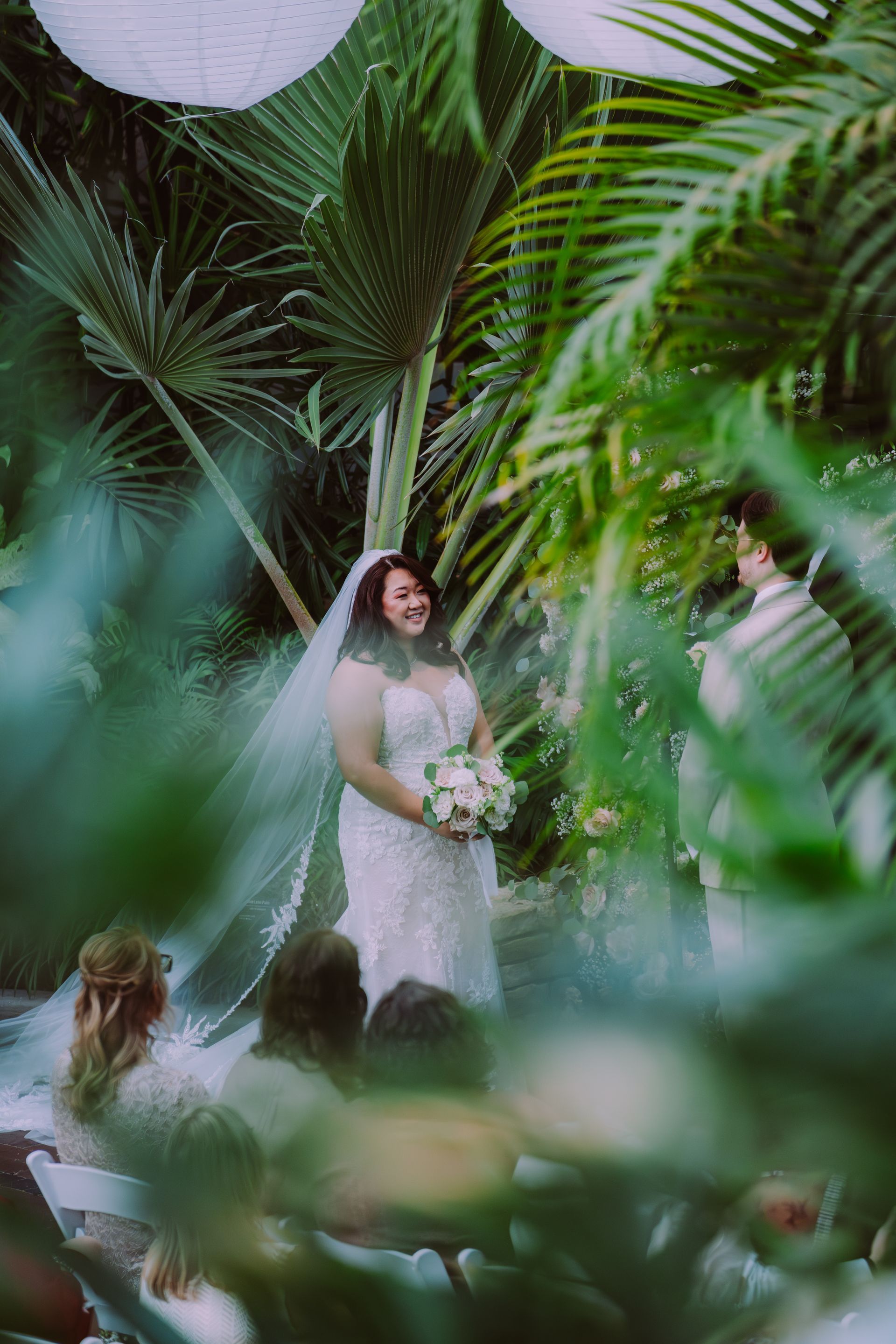 Bride in white dress holding bouquet, standing under palm leaves during wedding ceremony.
