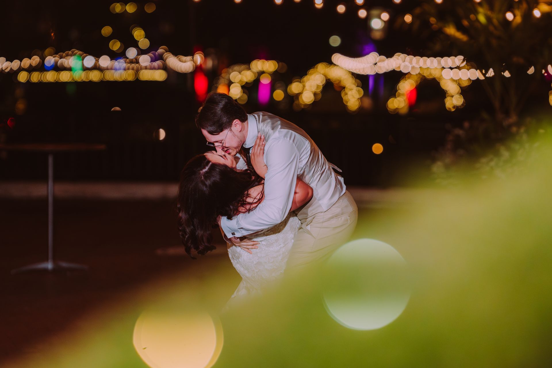 Couple kissing, he dips her; evening wedding, string lights.