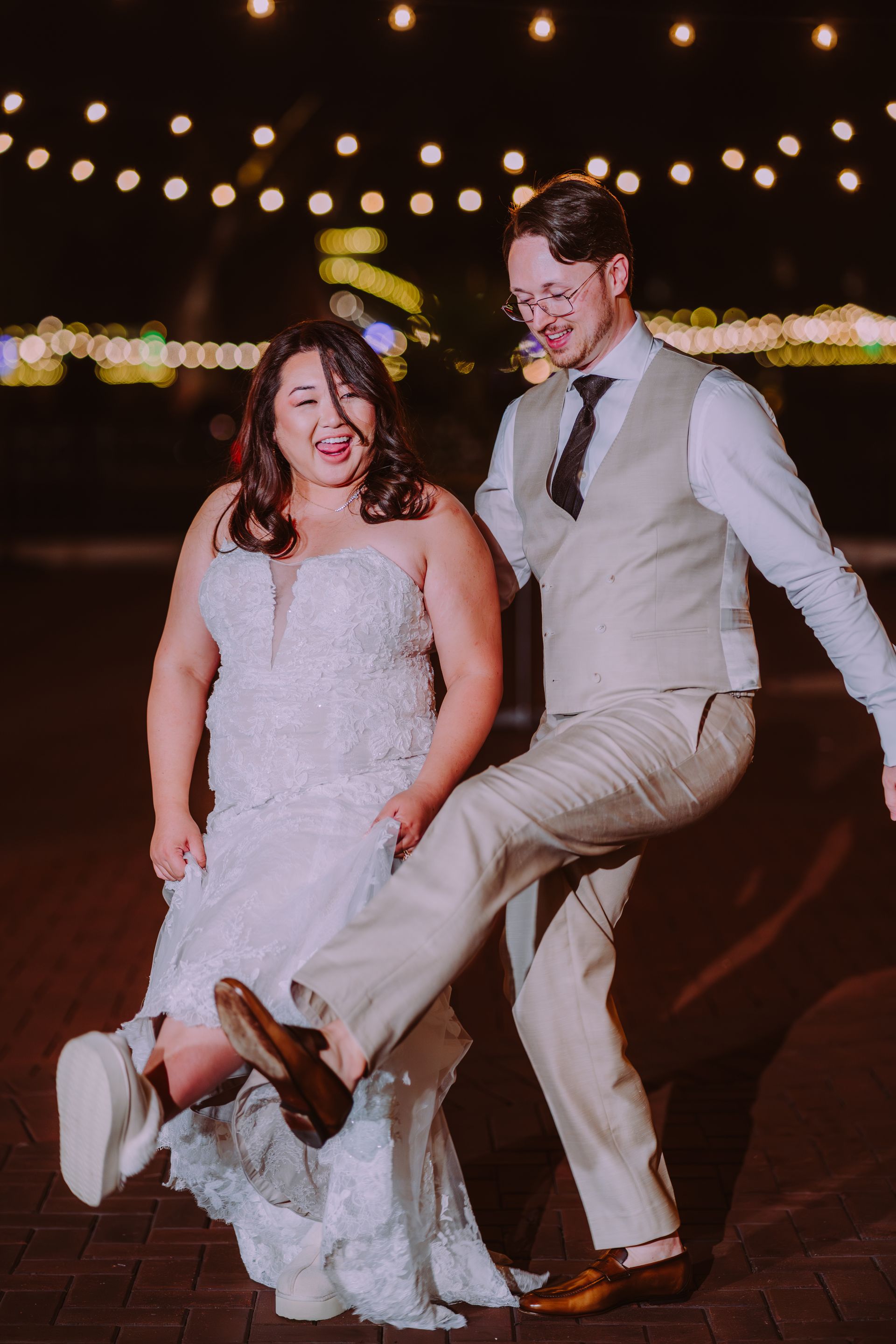 Newly married couple joyfully kicking, dressed in wedding attire at an outdoor evening reception, with string lights.