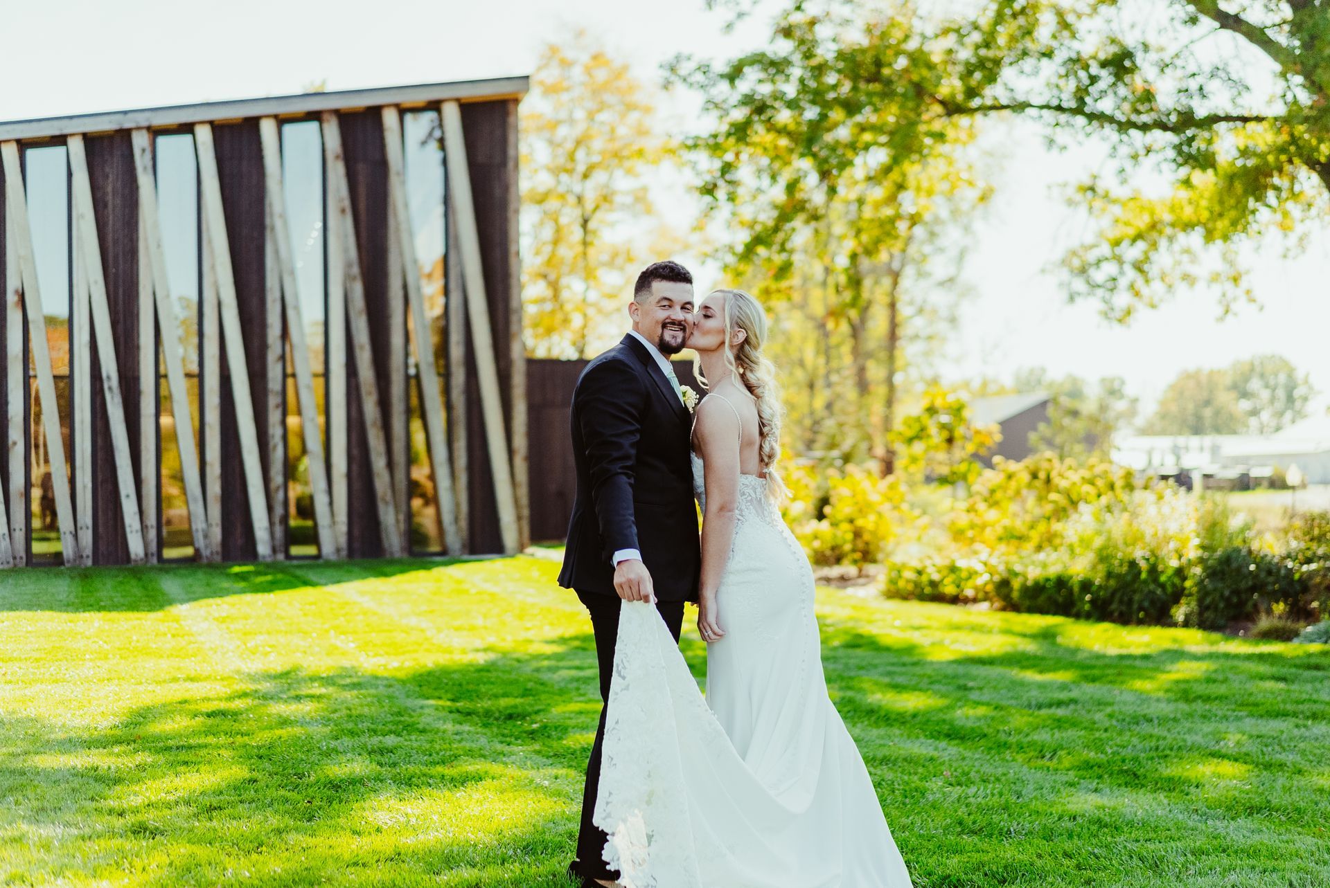 A bride and groom are kissing in the grass in front of a wooden structure.