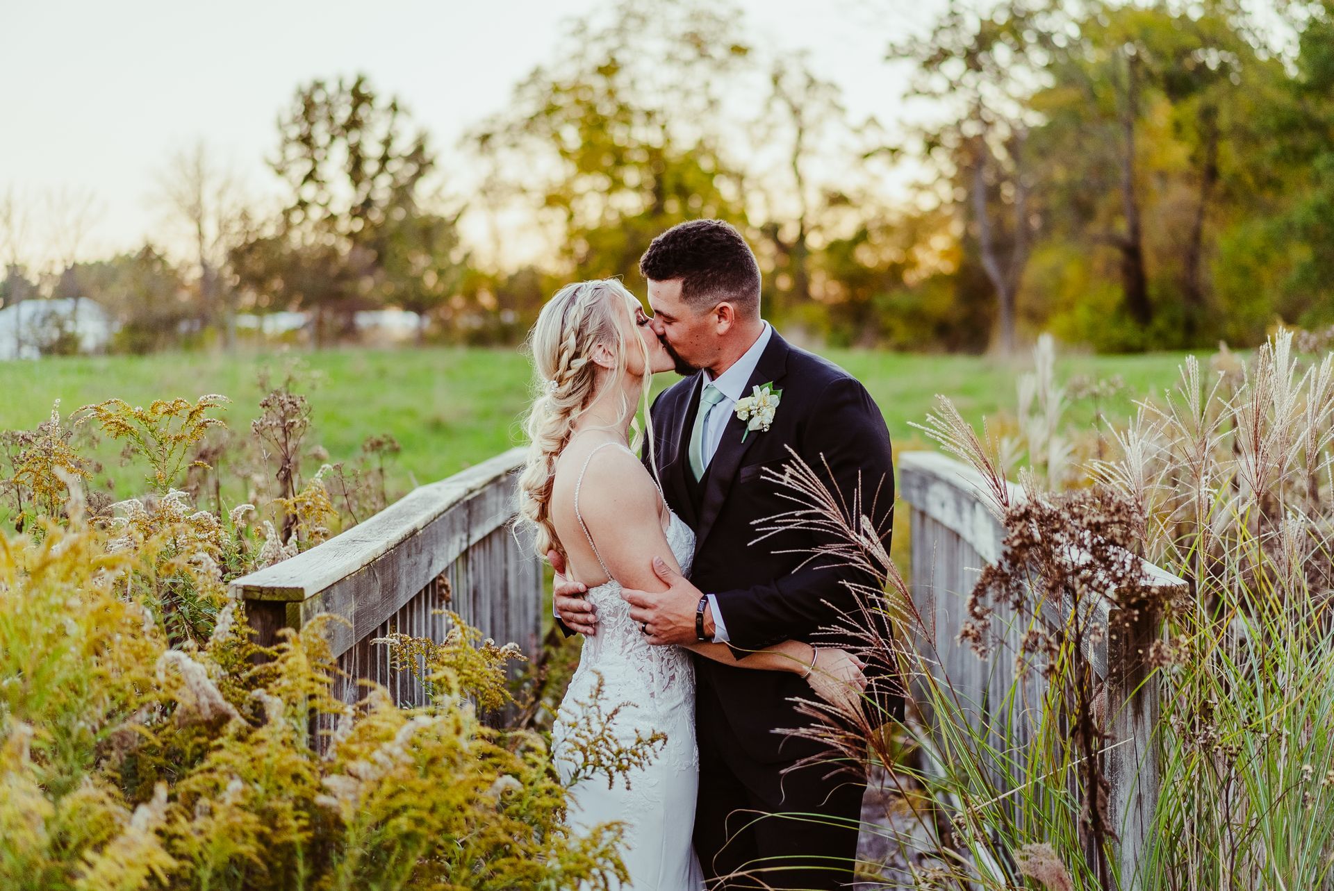 A bride and groom are kissing on a wooden bridge in a field.