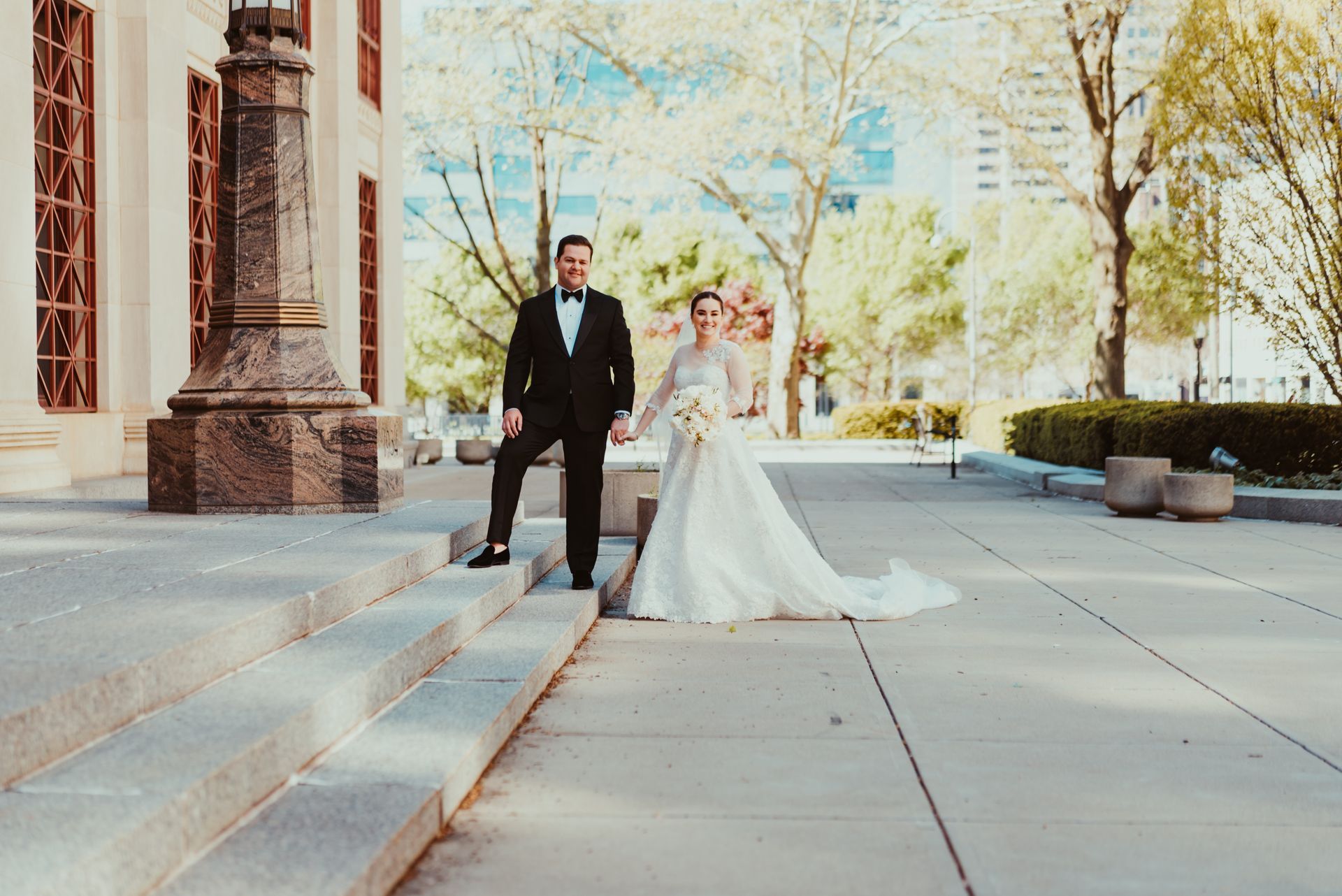 A bride and groom are standing on the steps of a building holding hands.