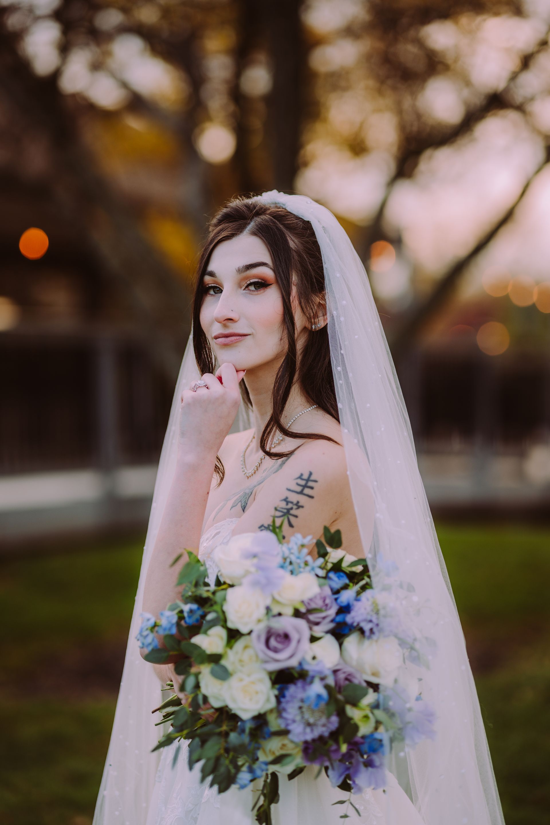 Bride with bouquet, veil, and arm tattoo, outdoors, holding her chin, smiling slightly.