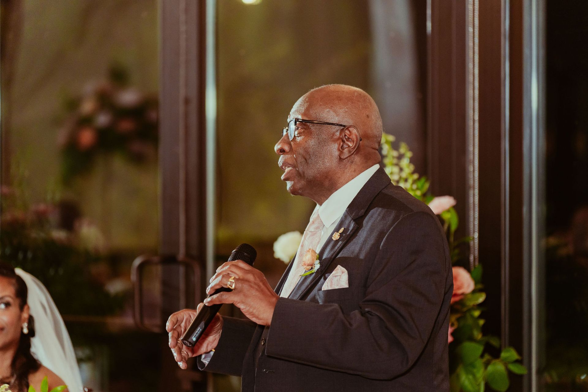 A man in a suit is holding a microphone and giving a speech at a wedding reception.