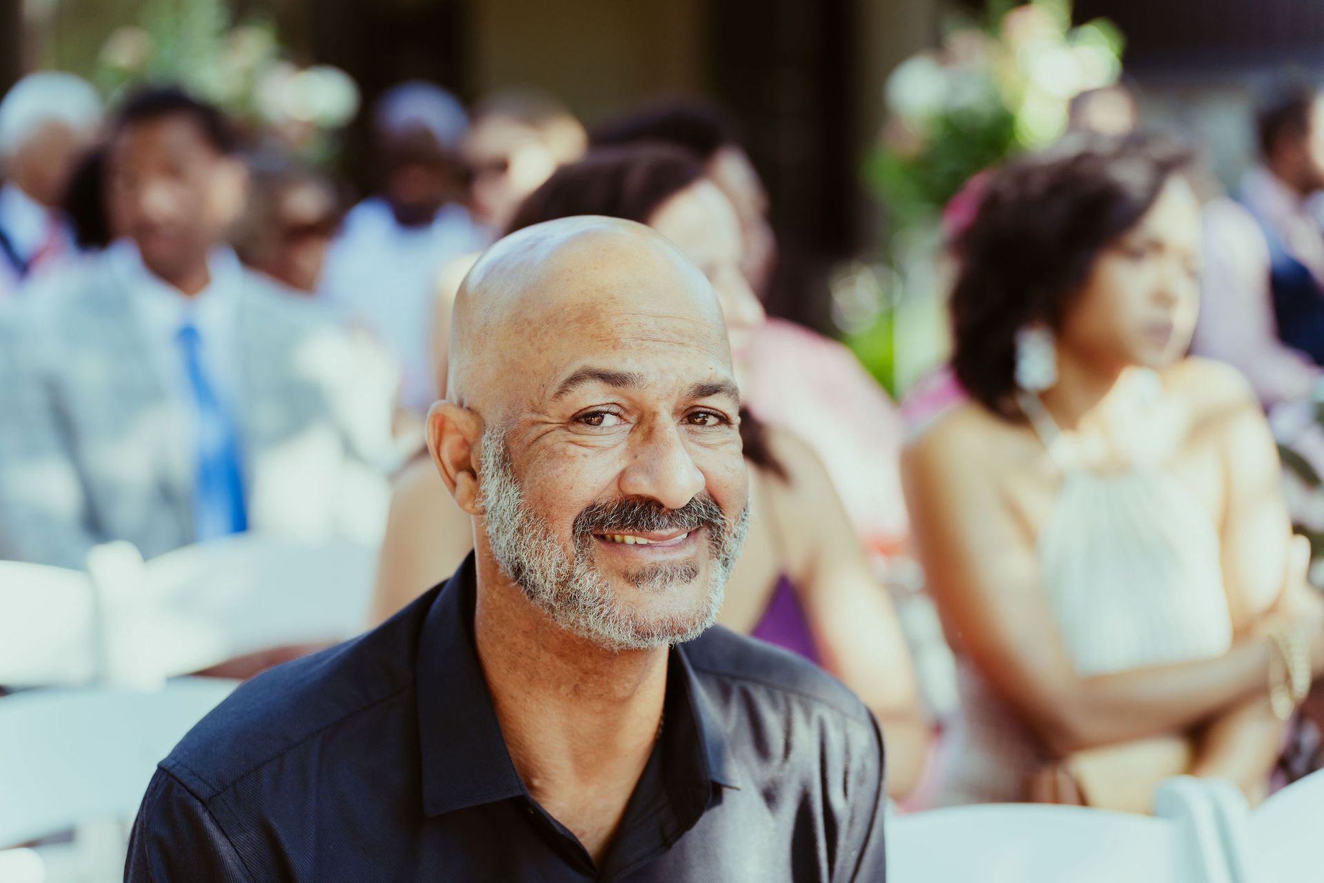 A man with a beard is smiling while sitting in a chair at a wedding.