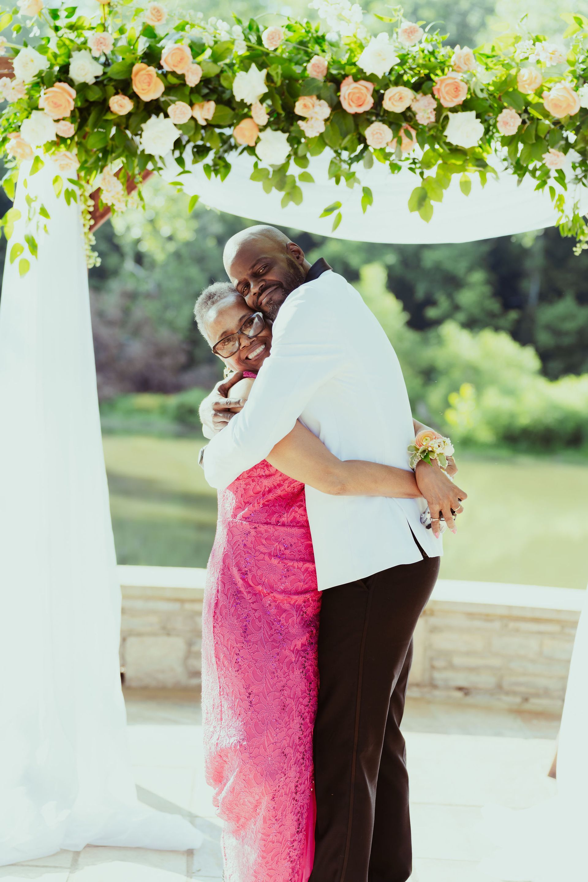 A man and woman are hugging under a floral arch . the woman is wearing a pink dress.