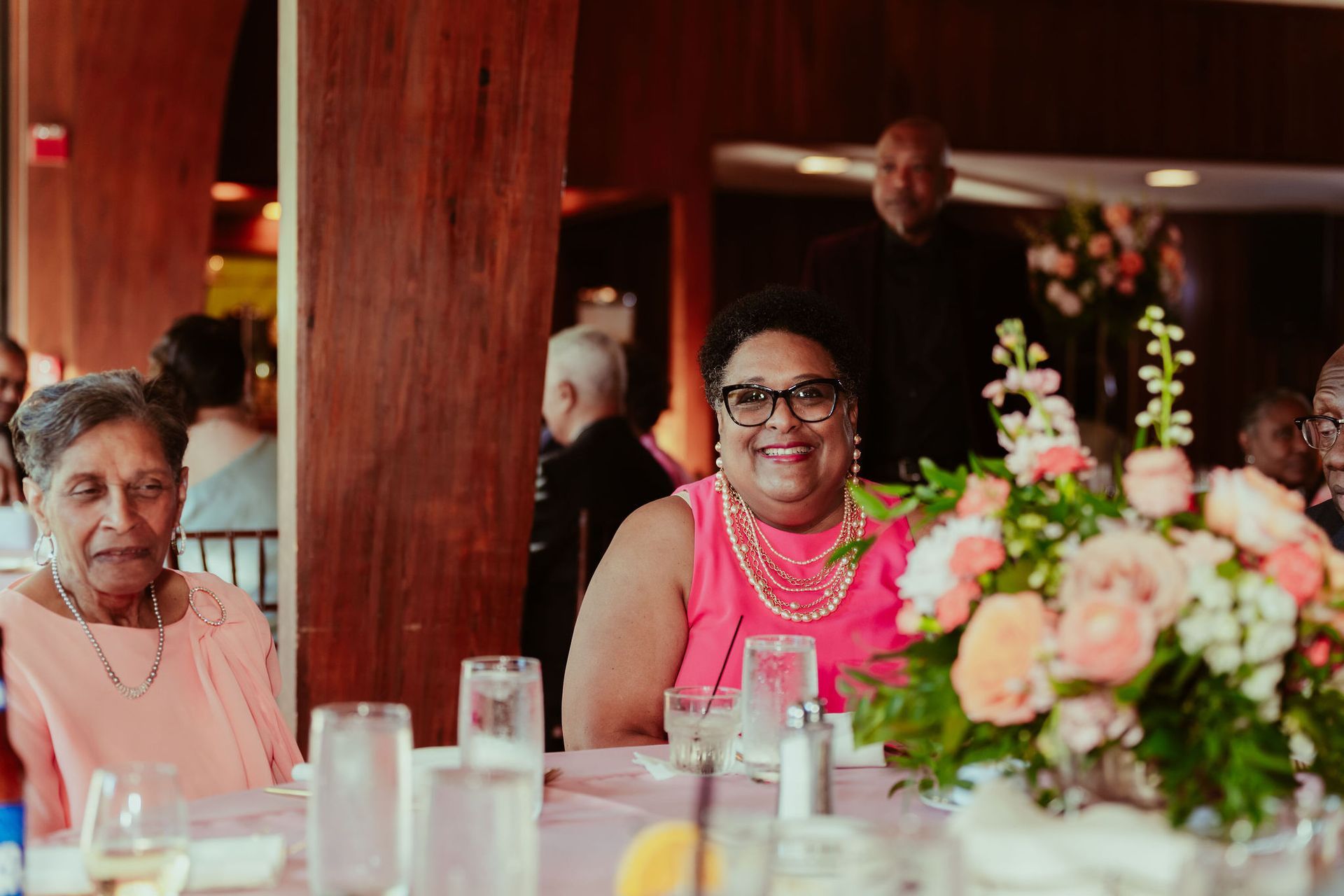 A woman in a pink dress is sitting at a table with a vase of flowers.