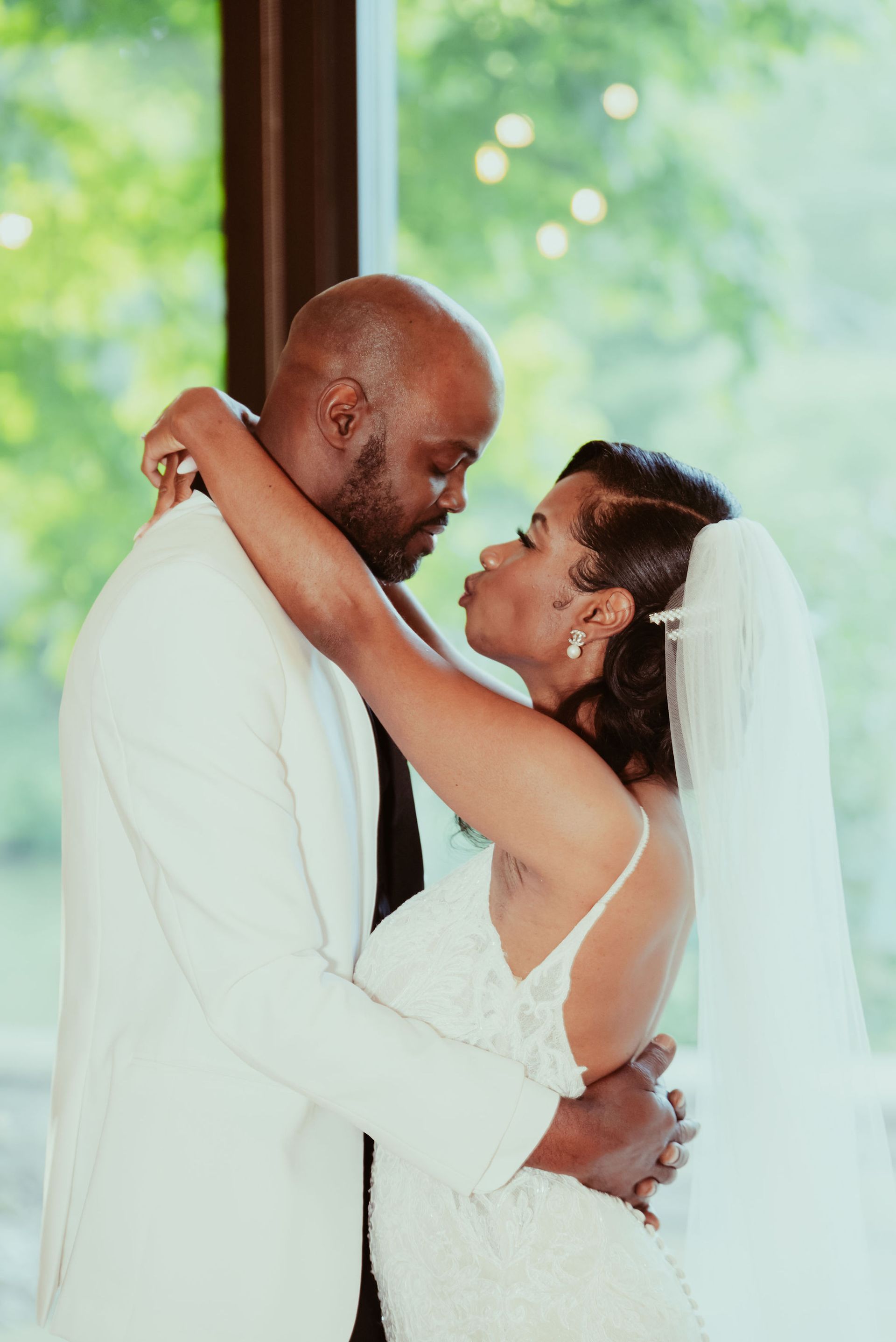 A bride and groom are hugging and kissing in front of a window.