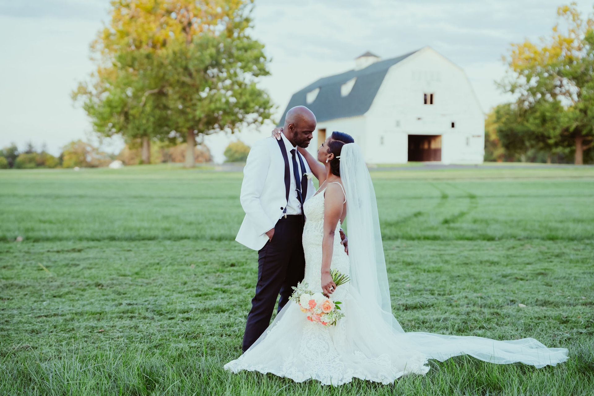 A bride and groom are standing in a field in front of a white barn.