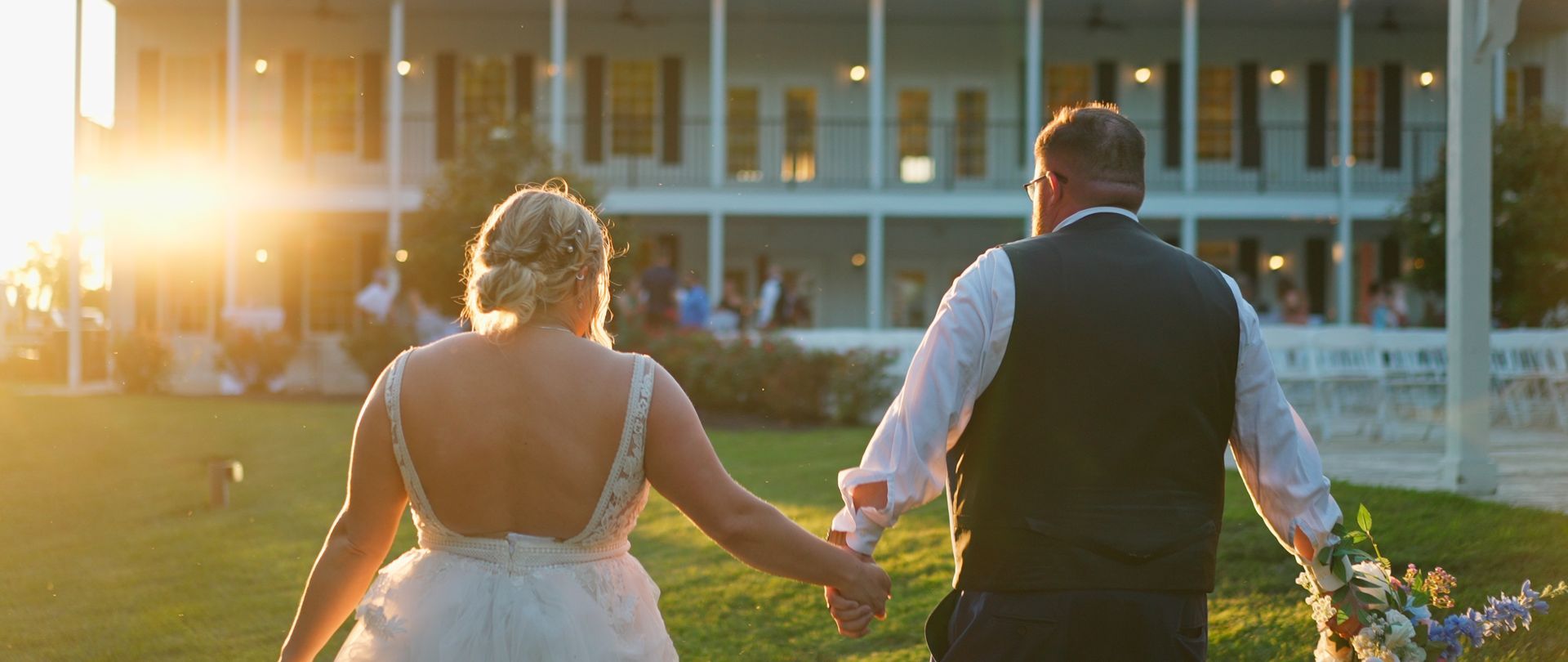 A bride and groom are walking in front of a large white house holding hands.