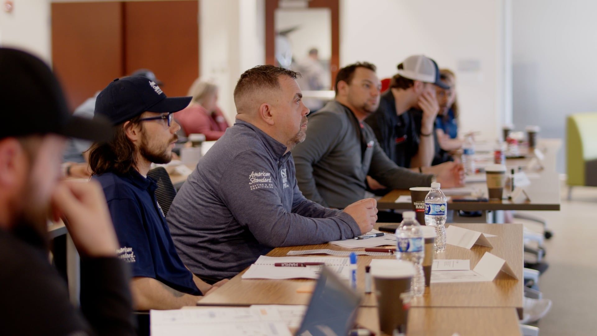 People seated at tables in a meeting room, listening attentively during a presentation.
