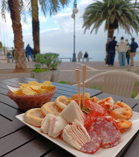 Piatto di antipasti con snack e patatine fritte su un tavolo, con vista sul lungomare con gente e palme.