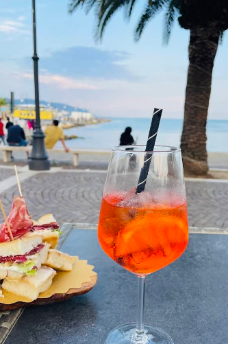 Cocktail e panini su un tavolo con vista sul mare, con una palma e persone sullo sfondo.