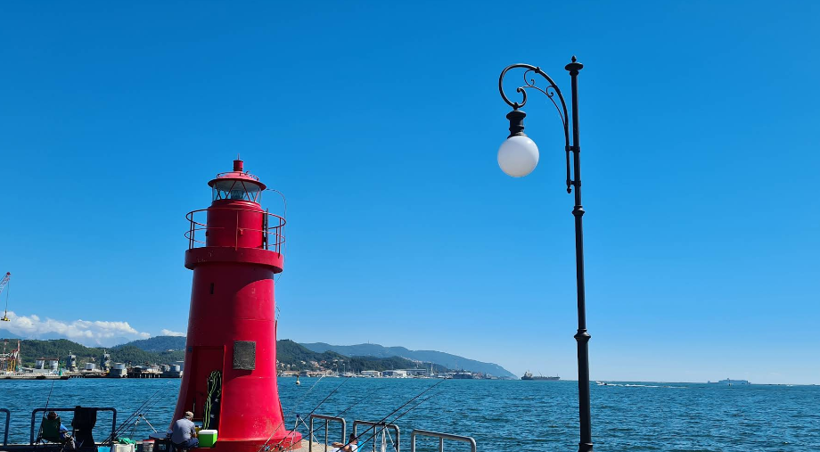 Un faro rosso e un lampione nero su un molo, sullo sfondo il cielo azzurro e il mare.