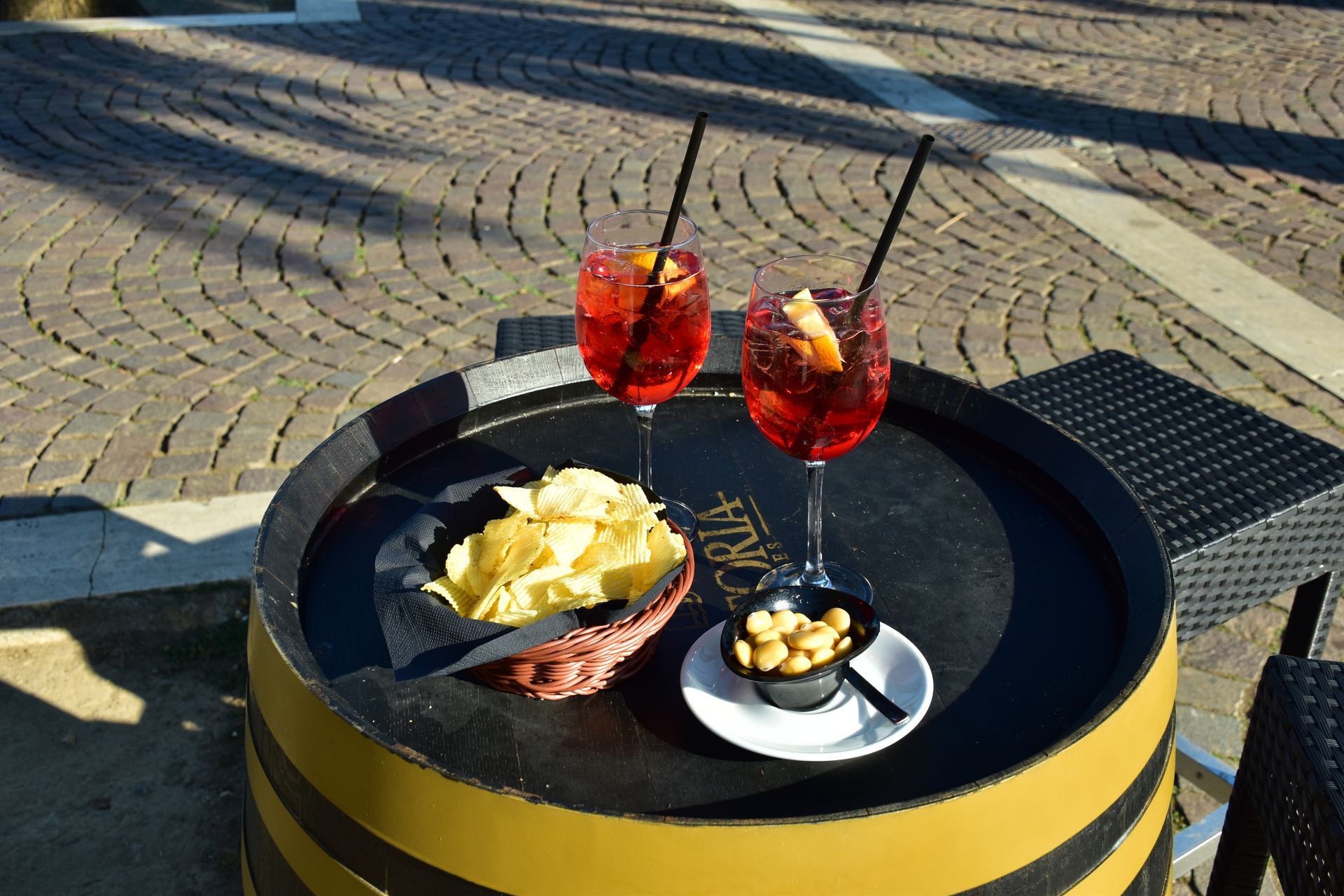 Due Aperol Spritz, patatine e noci su un tavolo a forma di botte all'aperto, con ciottoli e ombre.