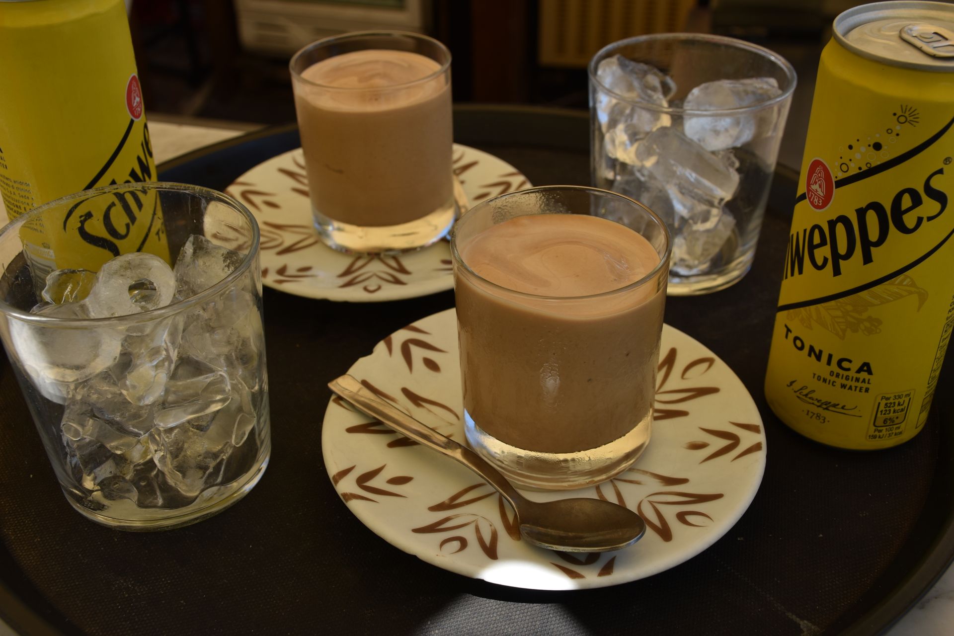 Bevande su un vassoio: due bicchieri di bevanda al cioccolato, due bicchieri di ghiaccio e due lattine gialle di acqua tonica Schweppes.