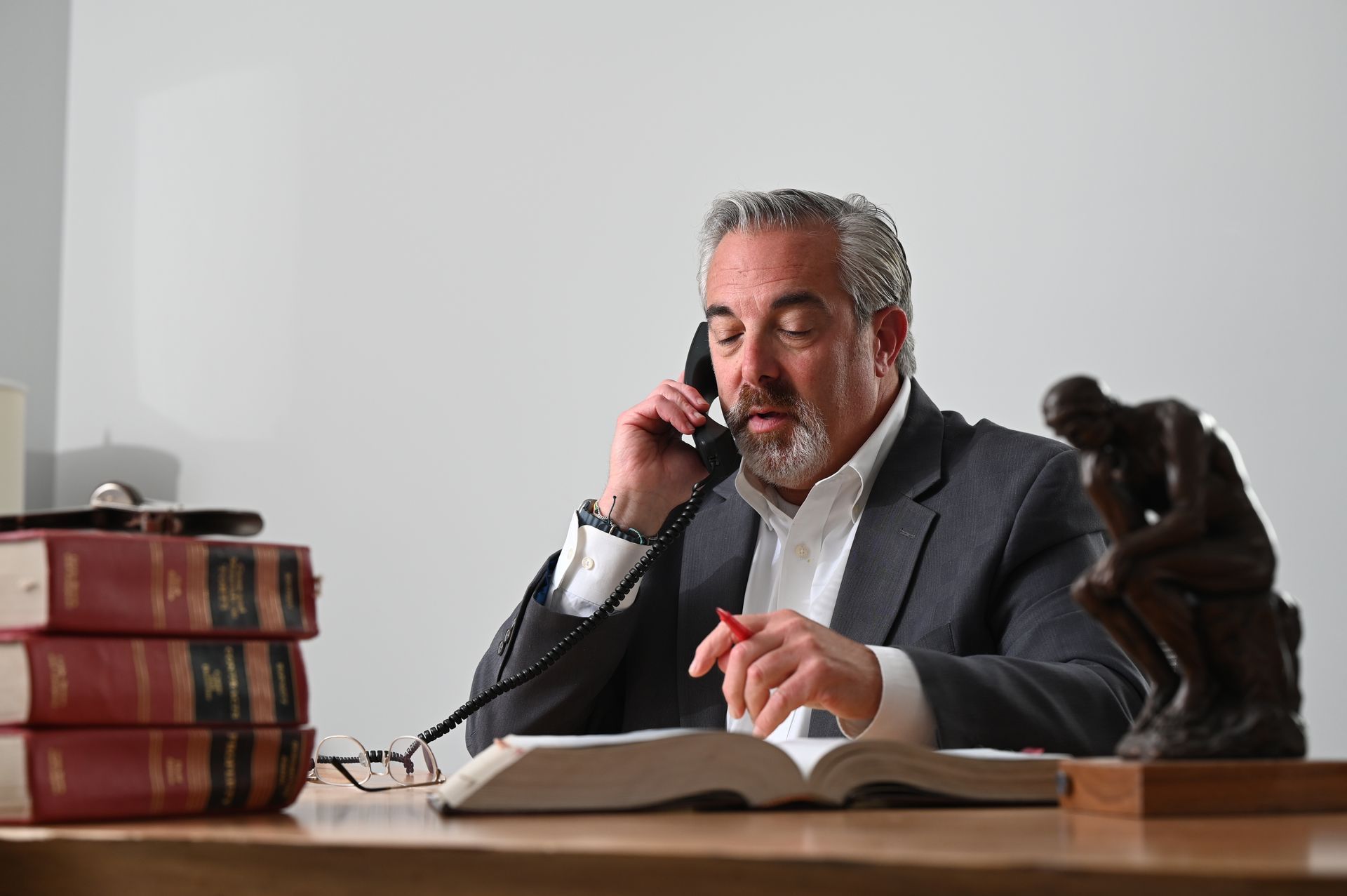 Attorney Robert Adelman in a gray suit on the phone, reading an open book at a desk beside stacked law books and a bronze statue