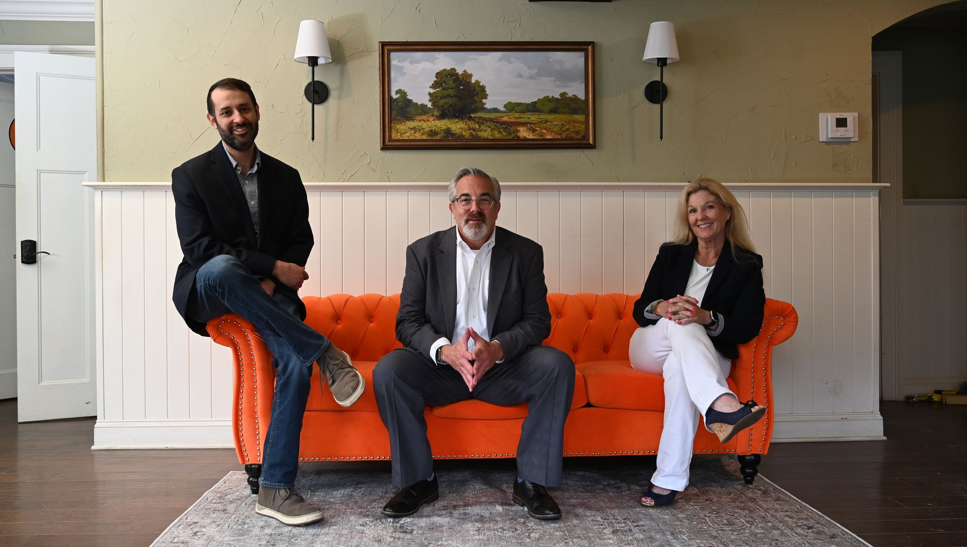 Three people seated on an orange couch in a living room, posing for a photo.