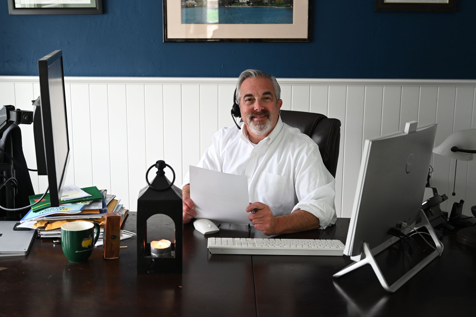 Man at a desk in an office, smiling and holding papers beside a computer and lantern-style lamp