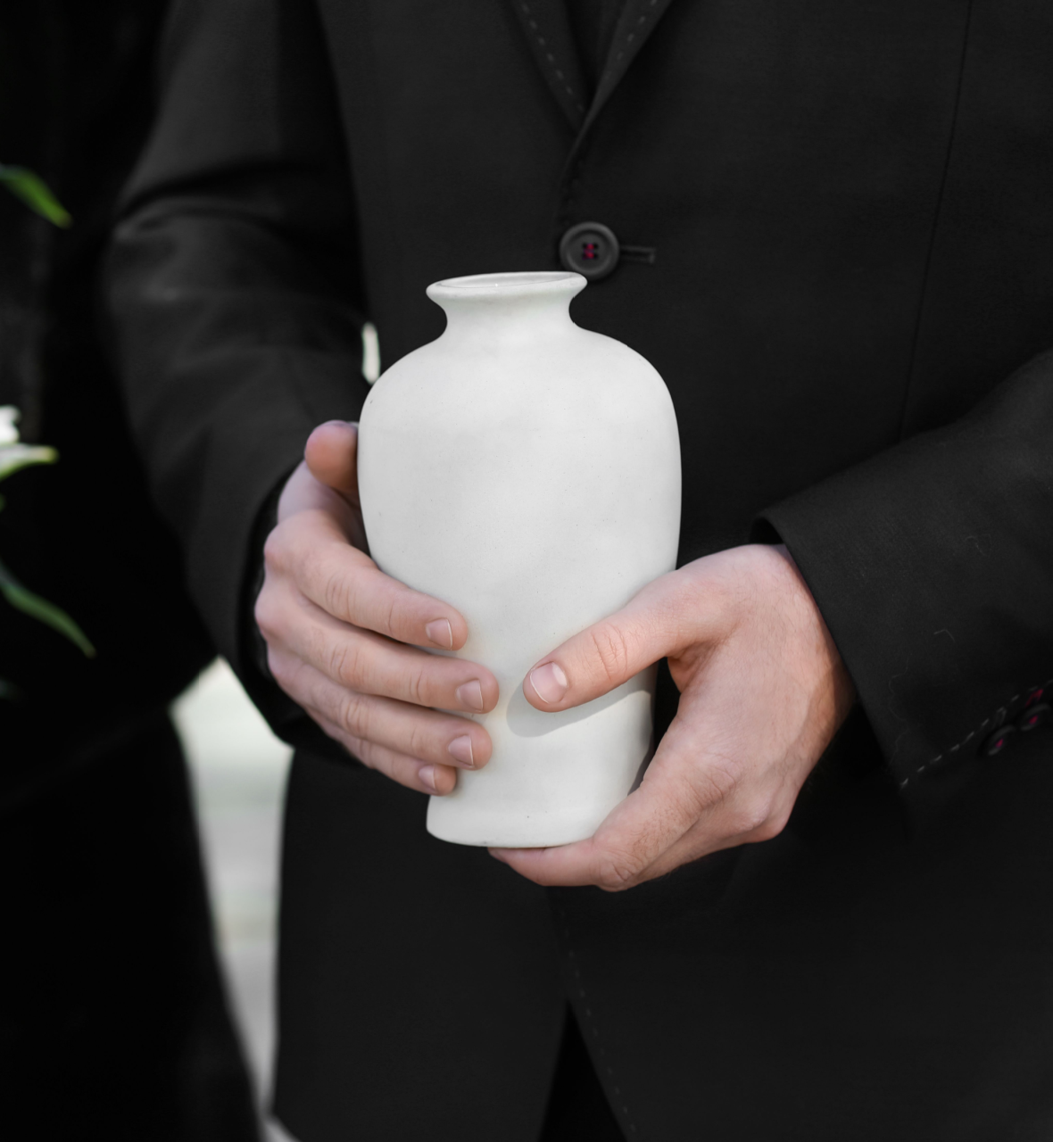 Person in black suit holding a white cremation urn, at a memorial.