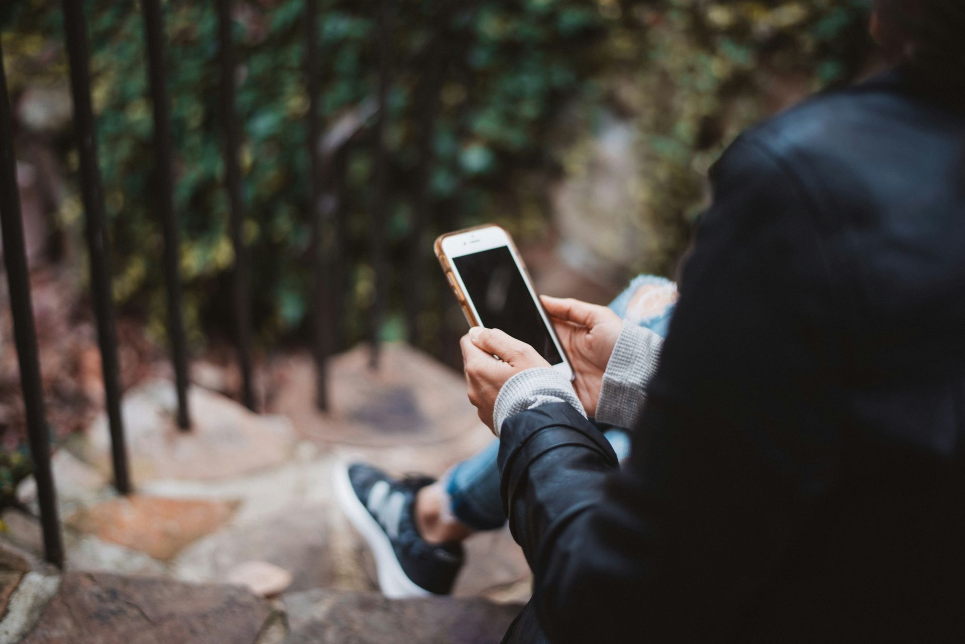 Person holding a phone, sitting on stone steps near greenery and black railings.