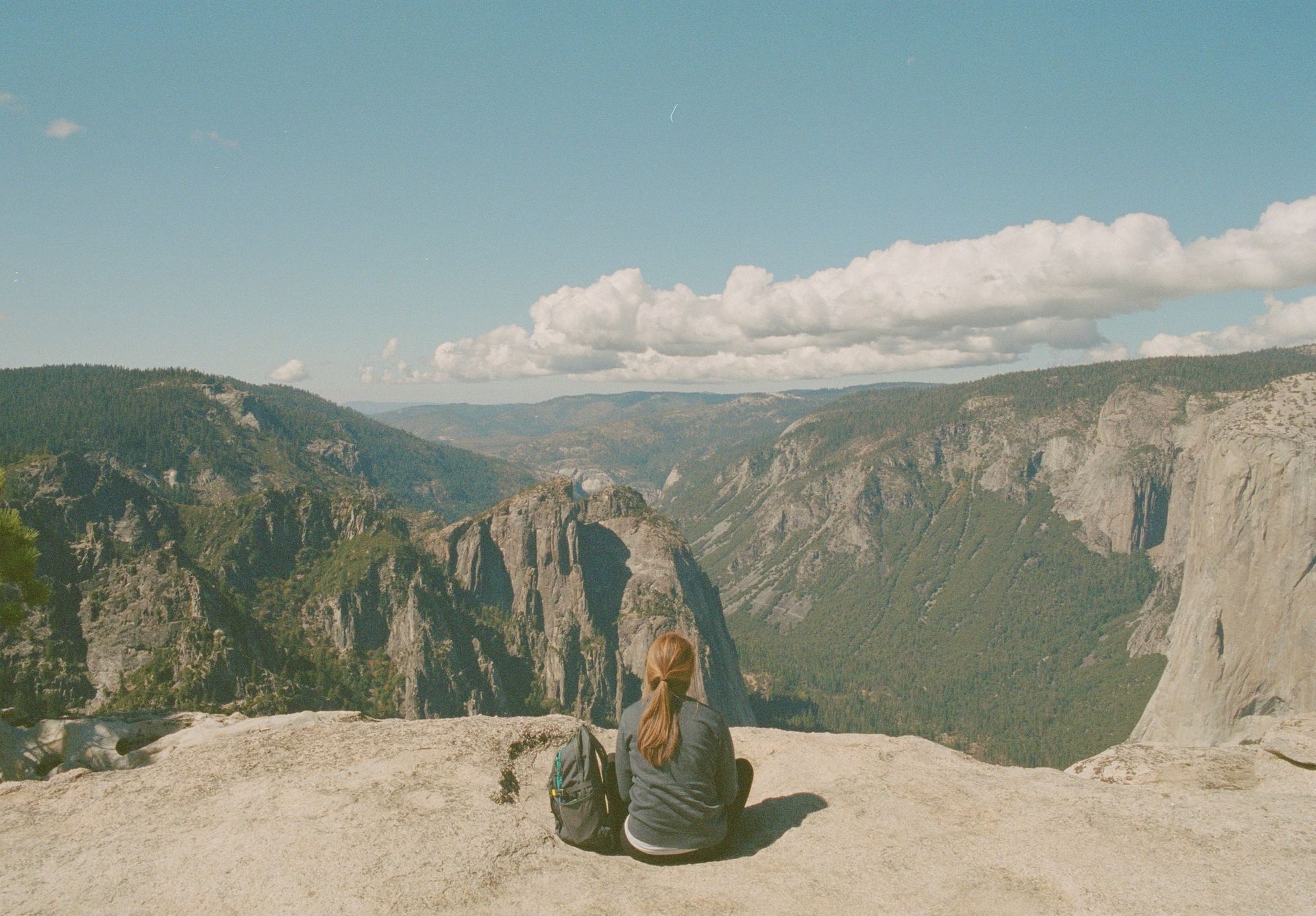 Woman seated on cliff edge, looking at vast mountain landscape under blue sky.