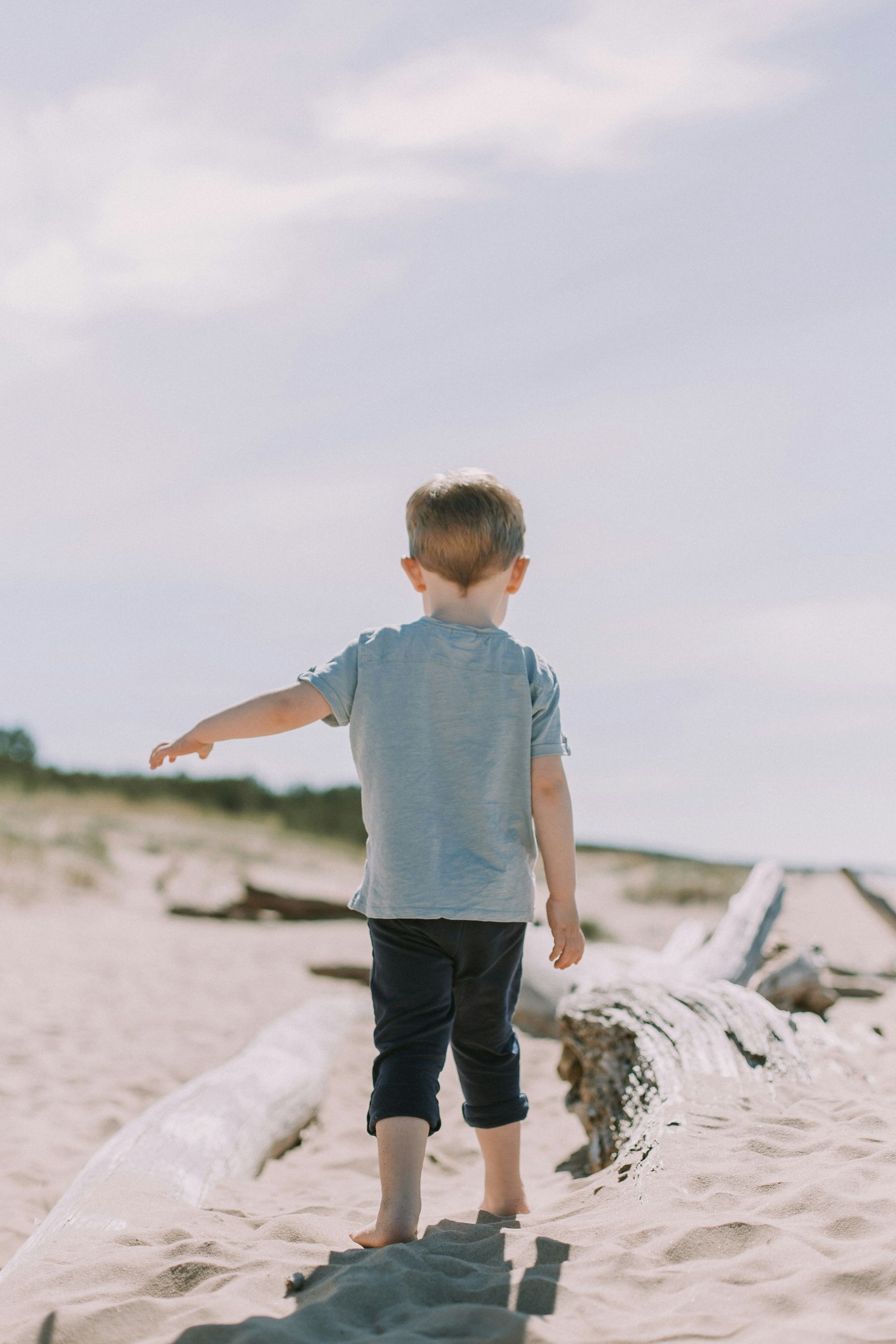 Young child in a gray shirt and dark pants stands on a sandy beach, arm outstretched.