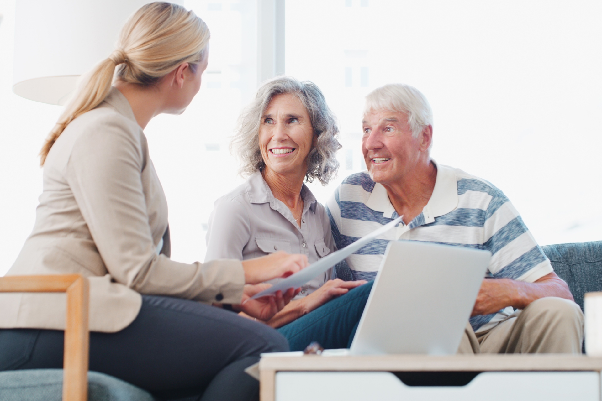 Woman advising a smiling older couple, reviewing documents. Laptop and table are present in a home setting.