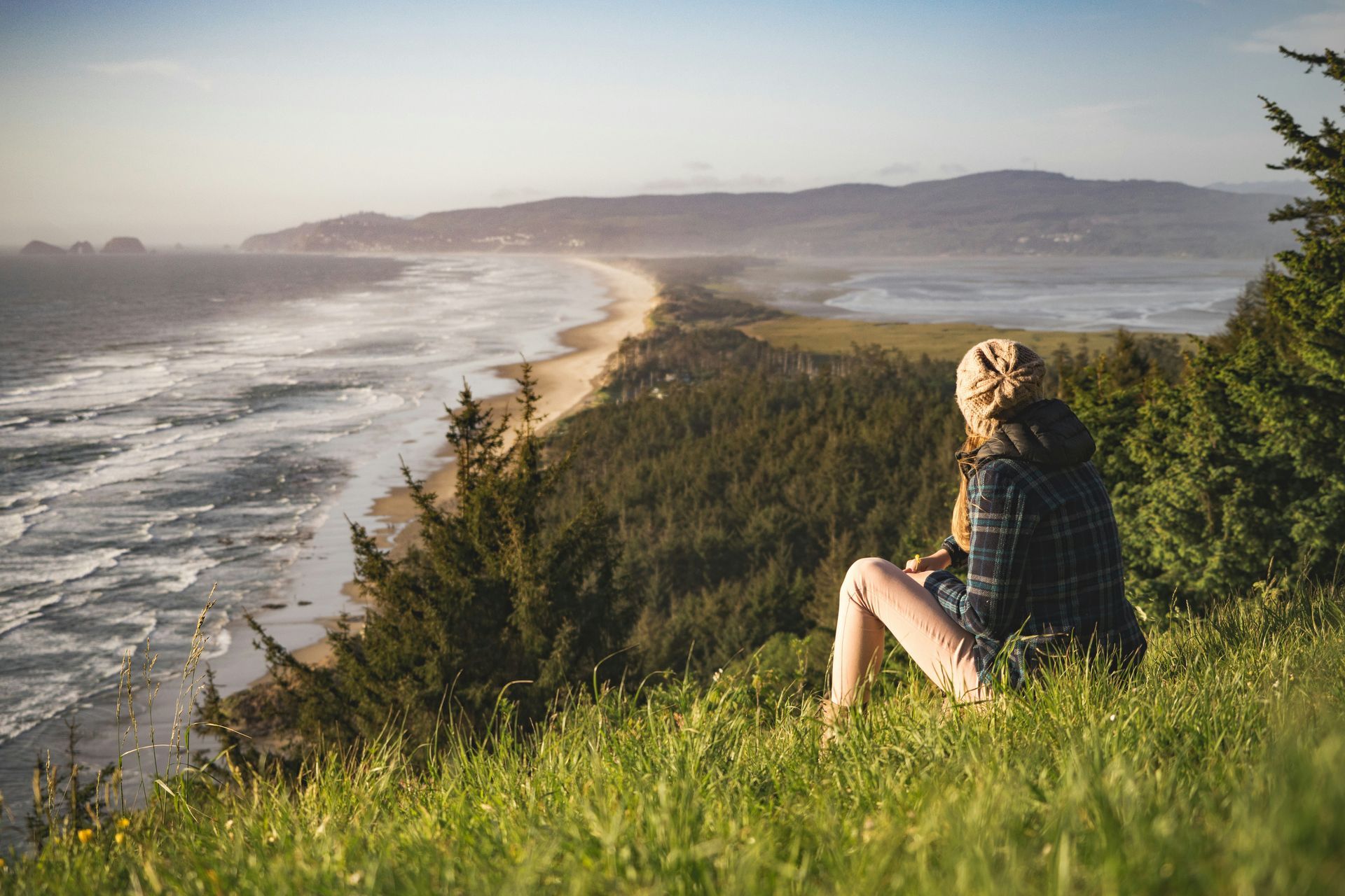 Person seated on a grassy hillside, looking at a coastal beach and ocean.