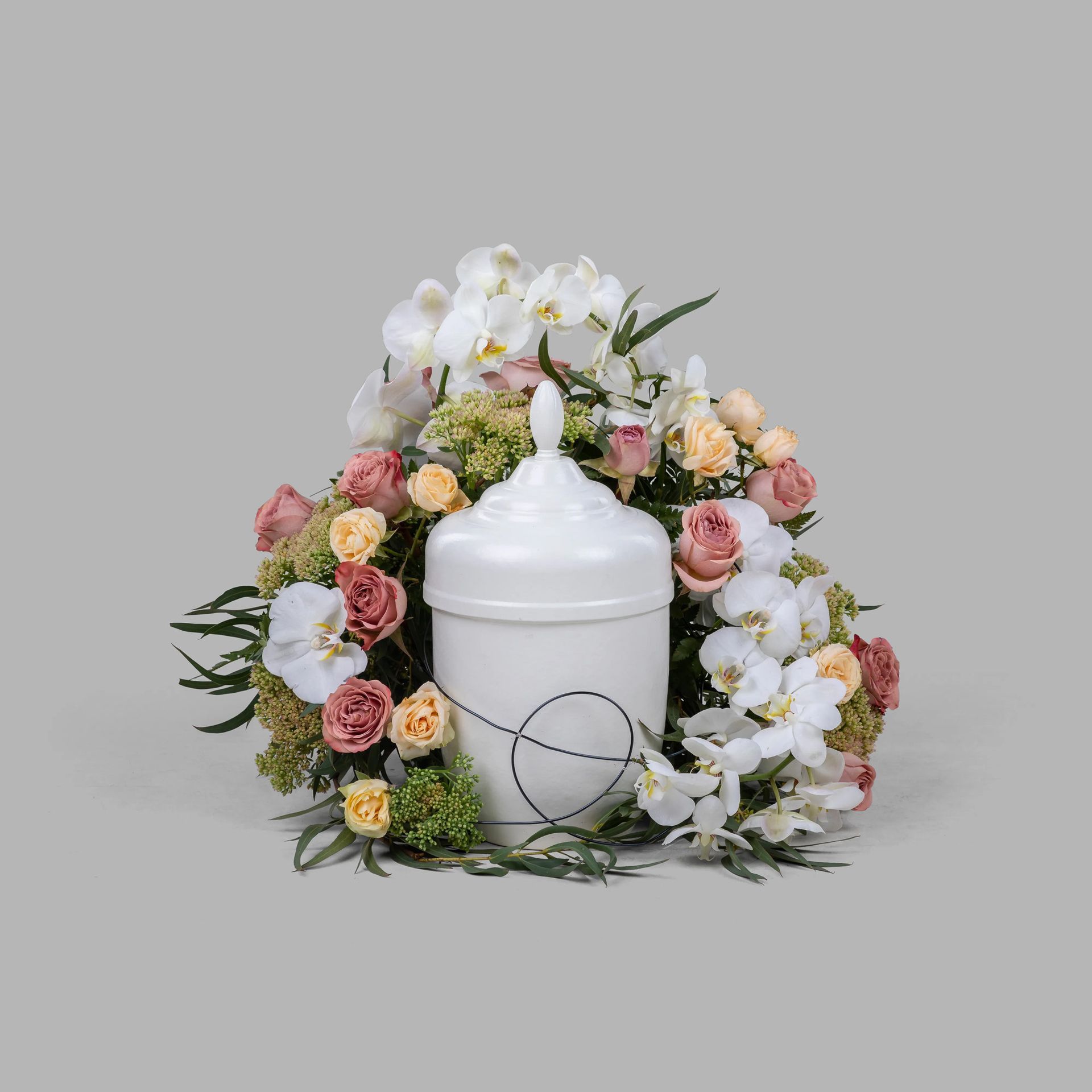 White cremation urn surrounded by floral arrangement: orchids, roses, greenery, against a gray background.