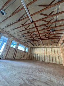 Interior of a room with exposed wooden beams, plywood floor, and spray foam insulation on the walls and ceiling.