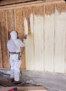 Person in protective suit spraying insulation foam onto a wooden wall in a building.