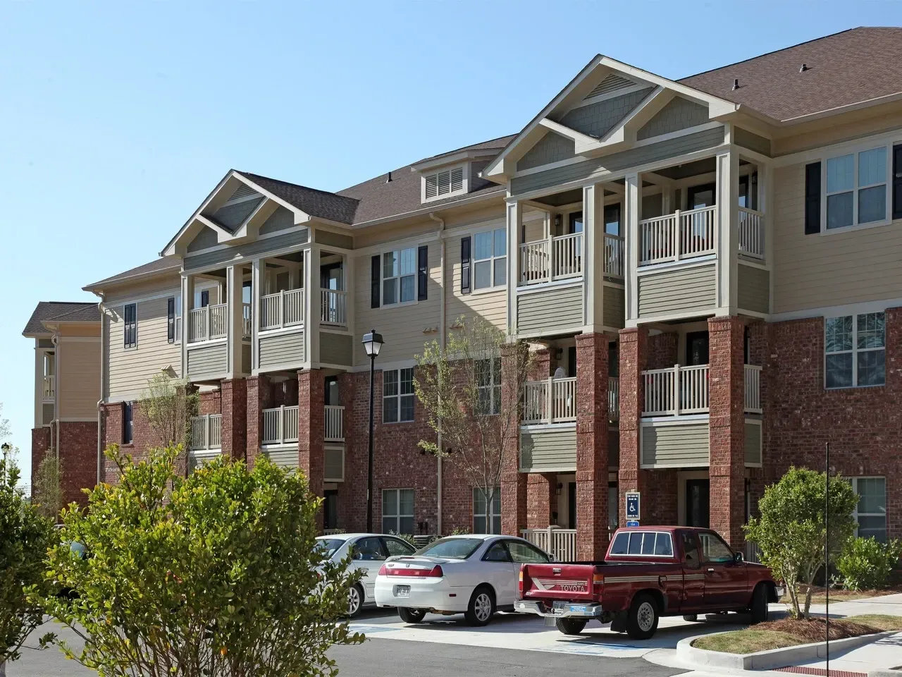 Exterior view of a multi-story apartment building with brick pillars, balconies, and a parking lot.