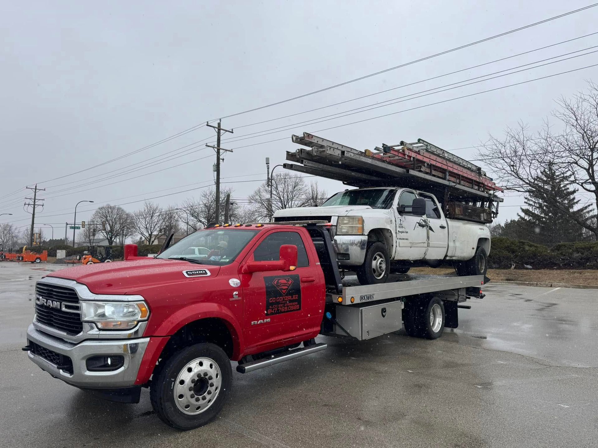 Red tow truck carrying a white pickup truck loaded with ladders on a cloudy day.