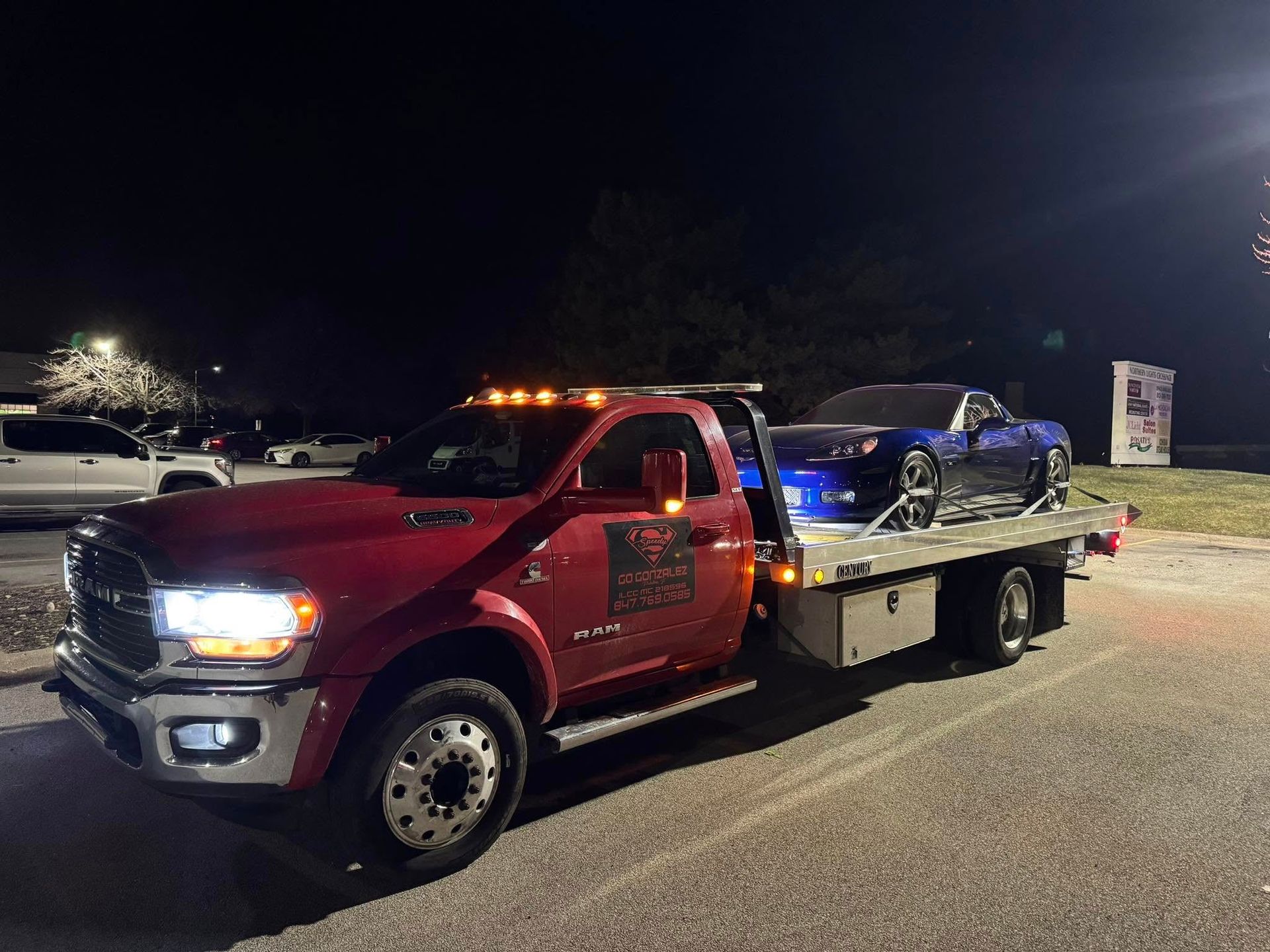 Red tow truck hauling a blue sports car at night; parking lot background.