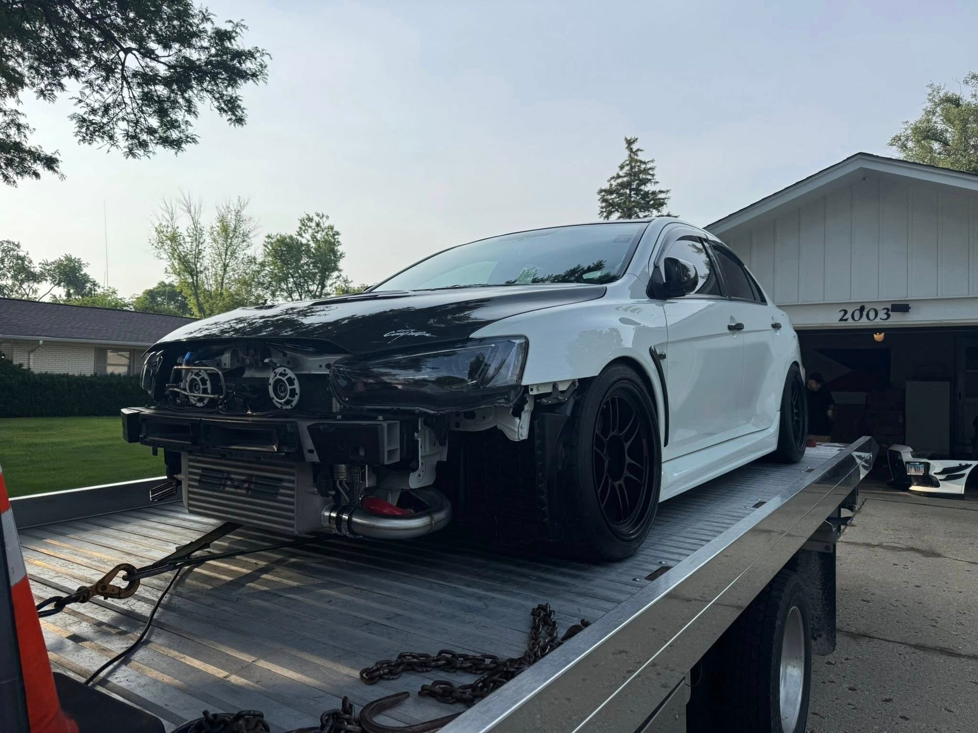 White Mitsubishi Evolution on a tow truck with front end damage, parked in front of a house.