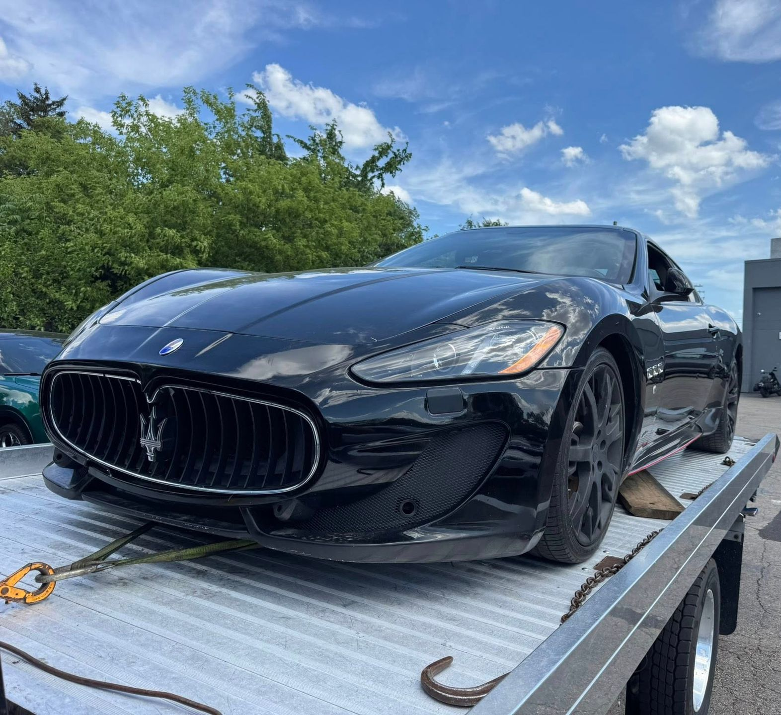 Black Maserati sports car on a tow truck bed under a bright, cloudy sky.
