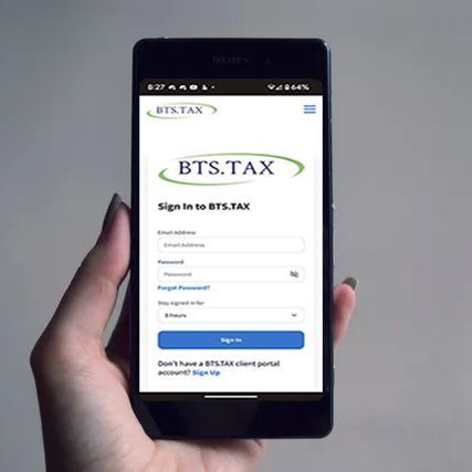 Hand holding a smartphone displaying the BTS.TAX login screen with fields for email and password.