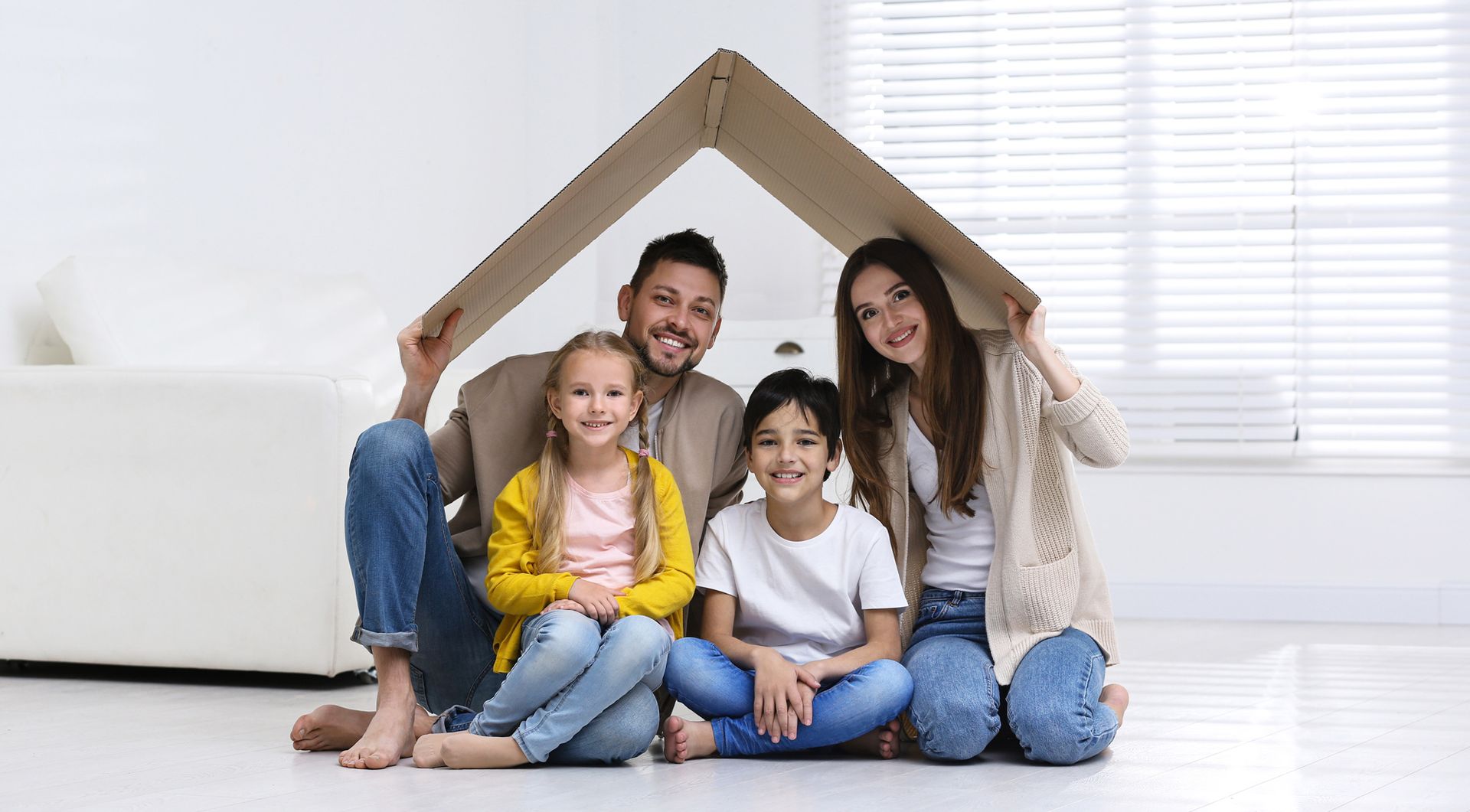 Family of four smiling under a cardboard house indoors.