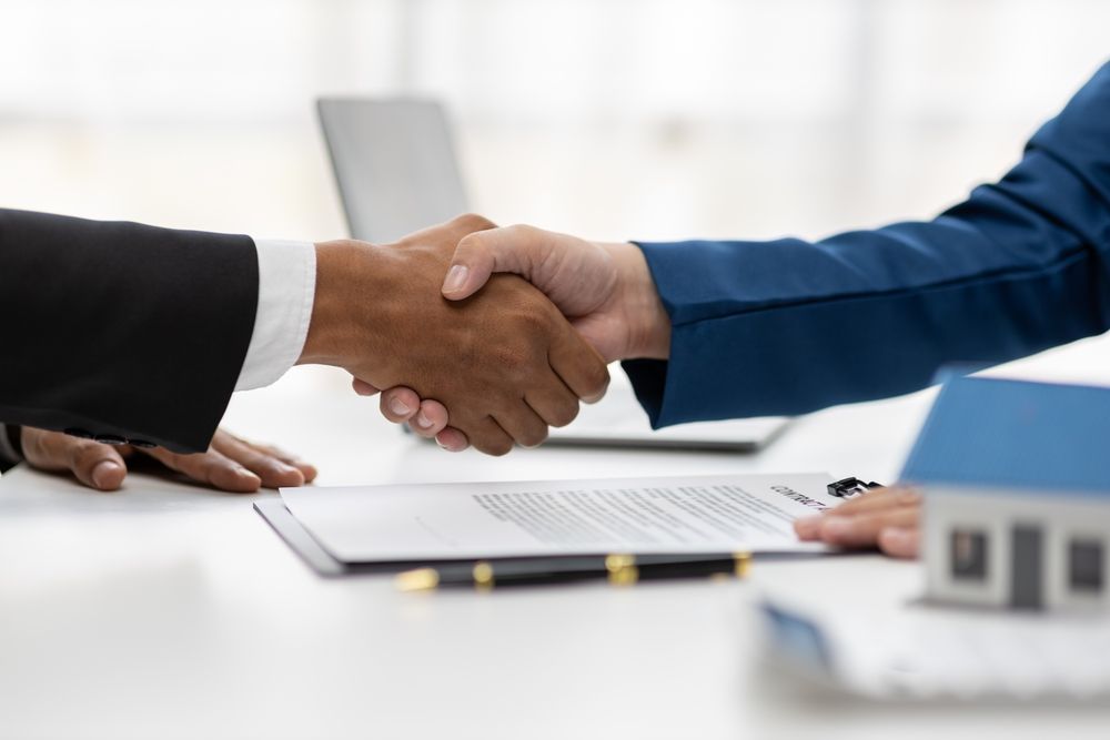 Two people shaking hands over a real estate contract on a table, small house model in the foreground.