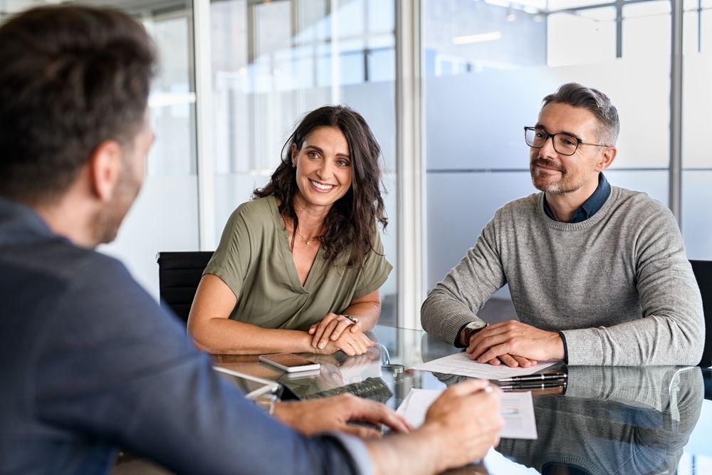Three people in a modern office meeting. Woman and man smiling at person opposite them, seated at a glass table.