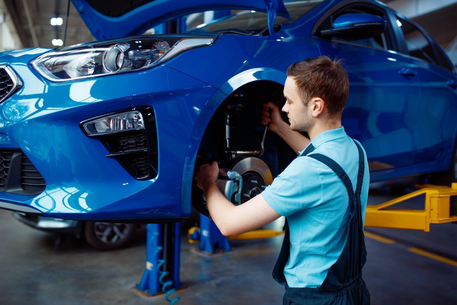 A mechanic in a blue uniform working on the front wheel assembly of a raised blue car inside a workshop.