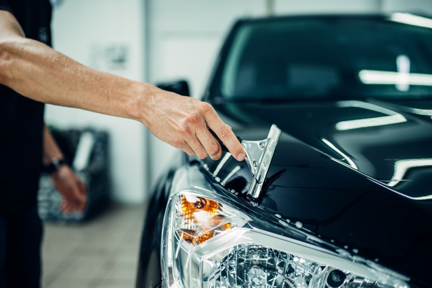 A hand uses a squeegee to smooth protective film onto the black hood of a car in a garage.