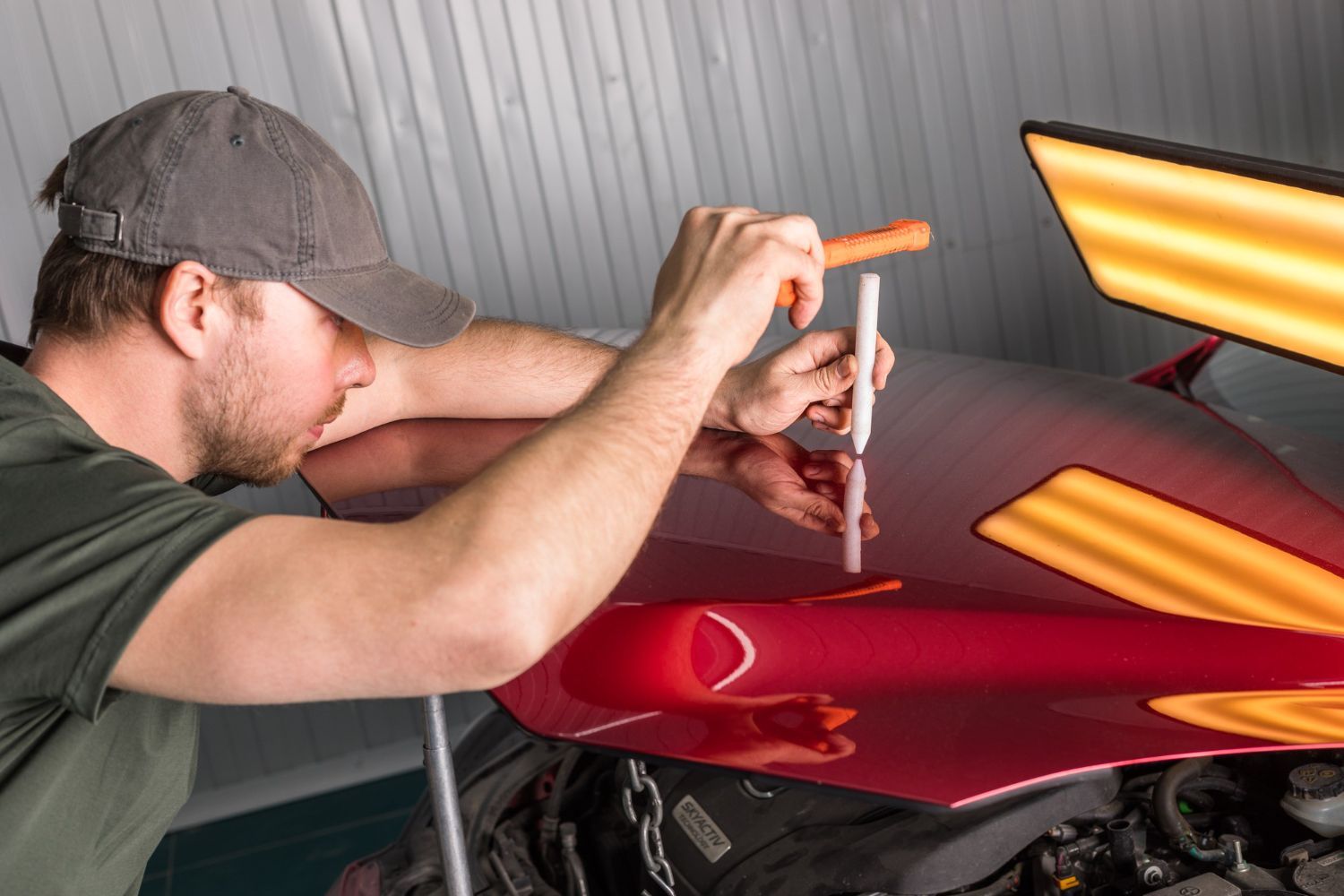 A technician uses a tool and a reflective light board to perform paintless dent repair on a red car hood.