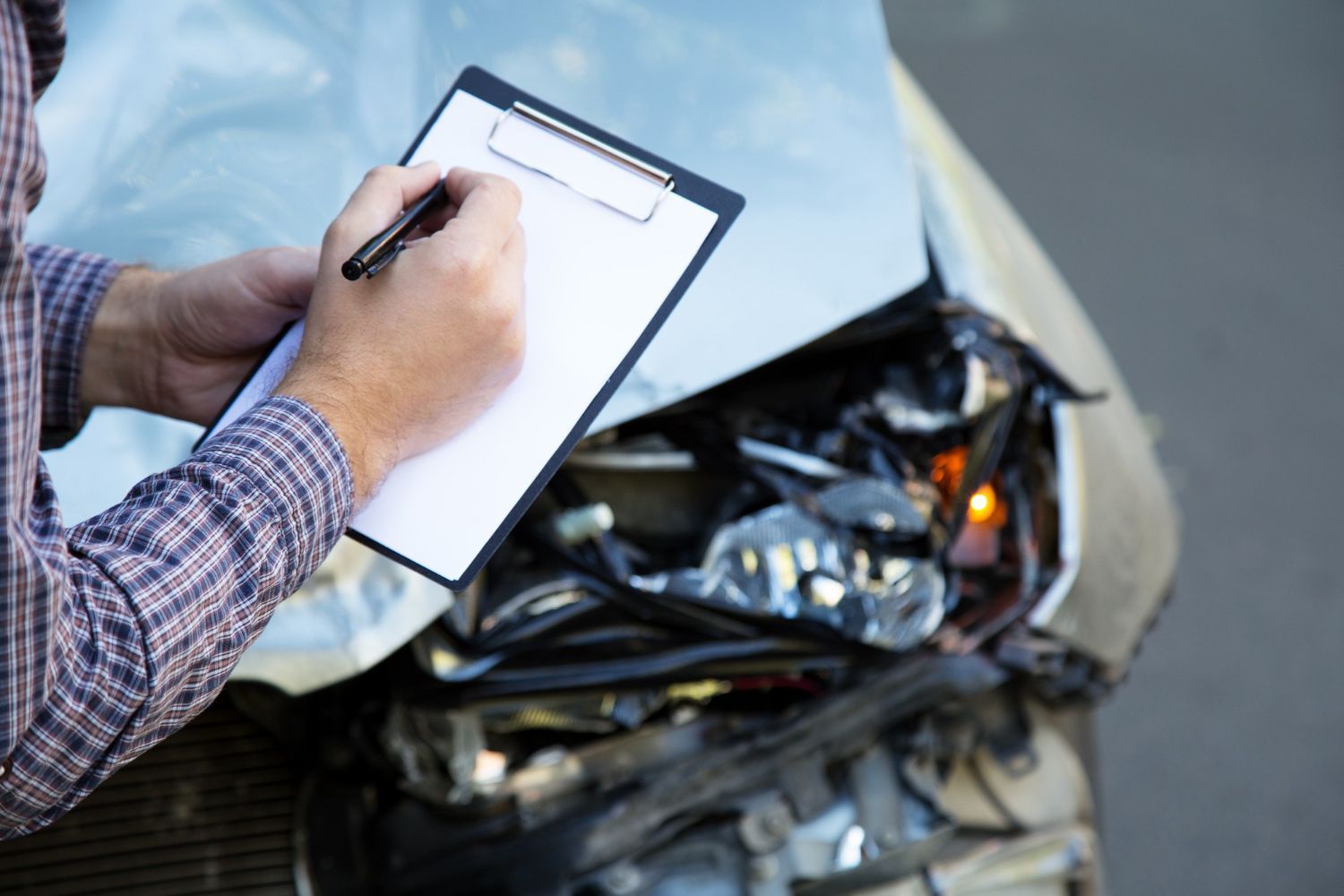 A person in a plaid shirt writes on a clipboard while standing next to a vehicle with significant front-end damage.