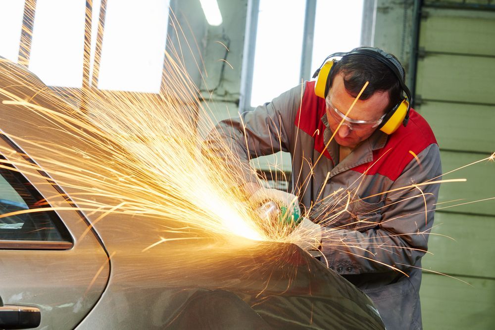 A person grinding a car fender, sparks flying. Wearing safety glasses, ear protection, and gloves.